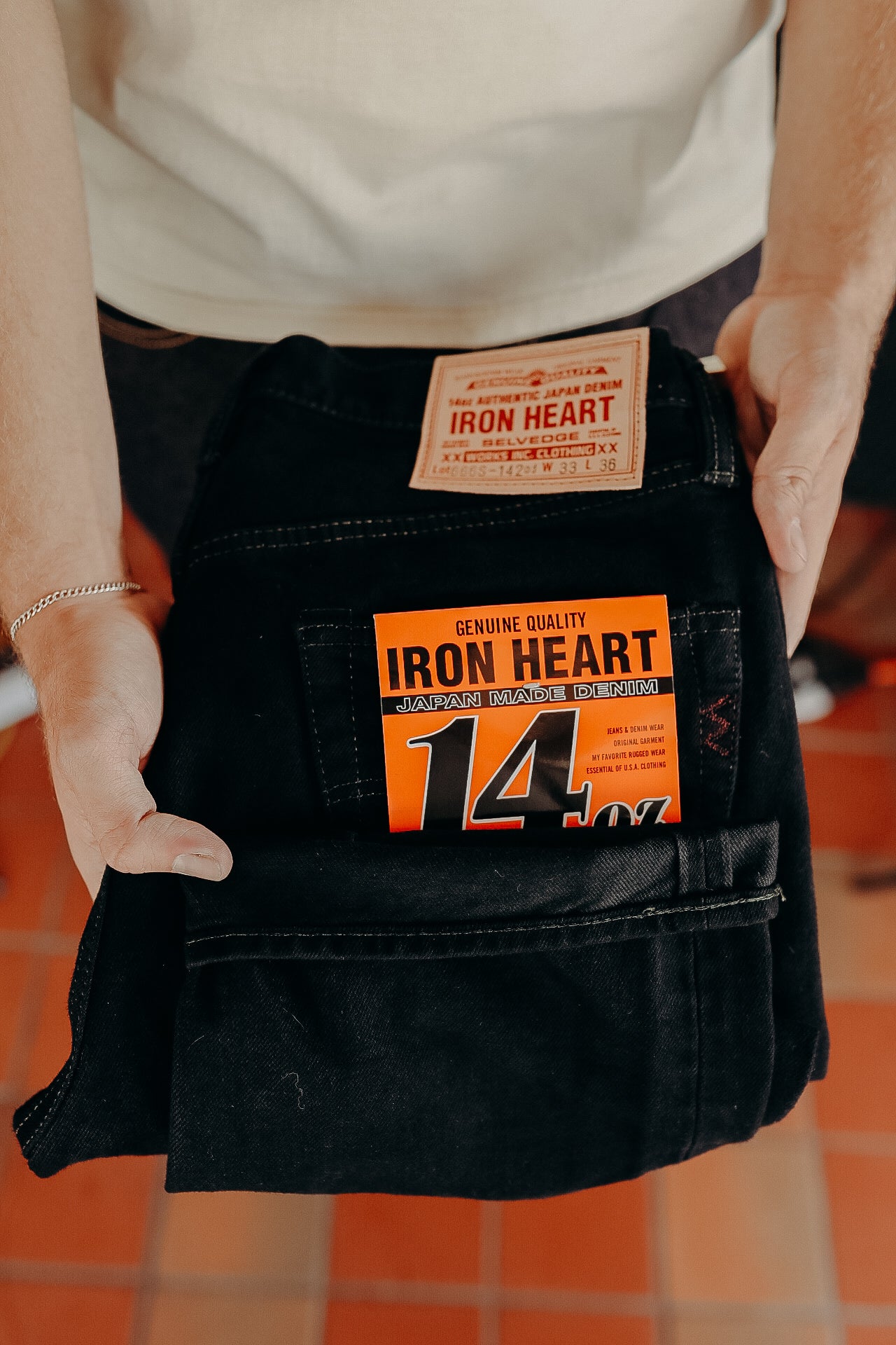Person holding folded black Iron Heart 14 oz Japan made denim jeans with orange label on tiled floor