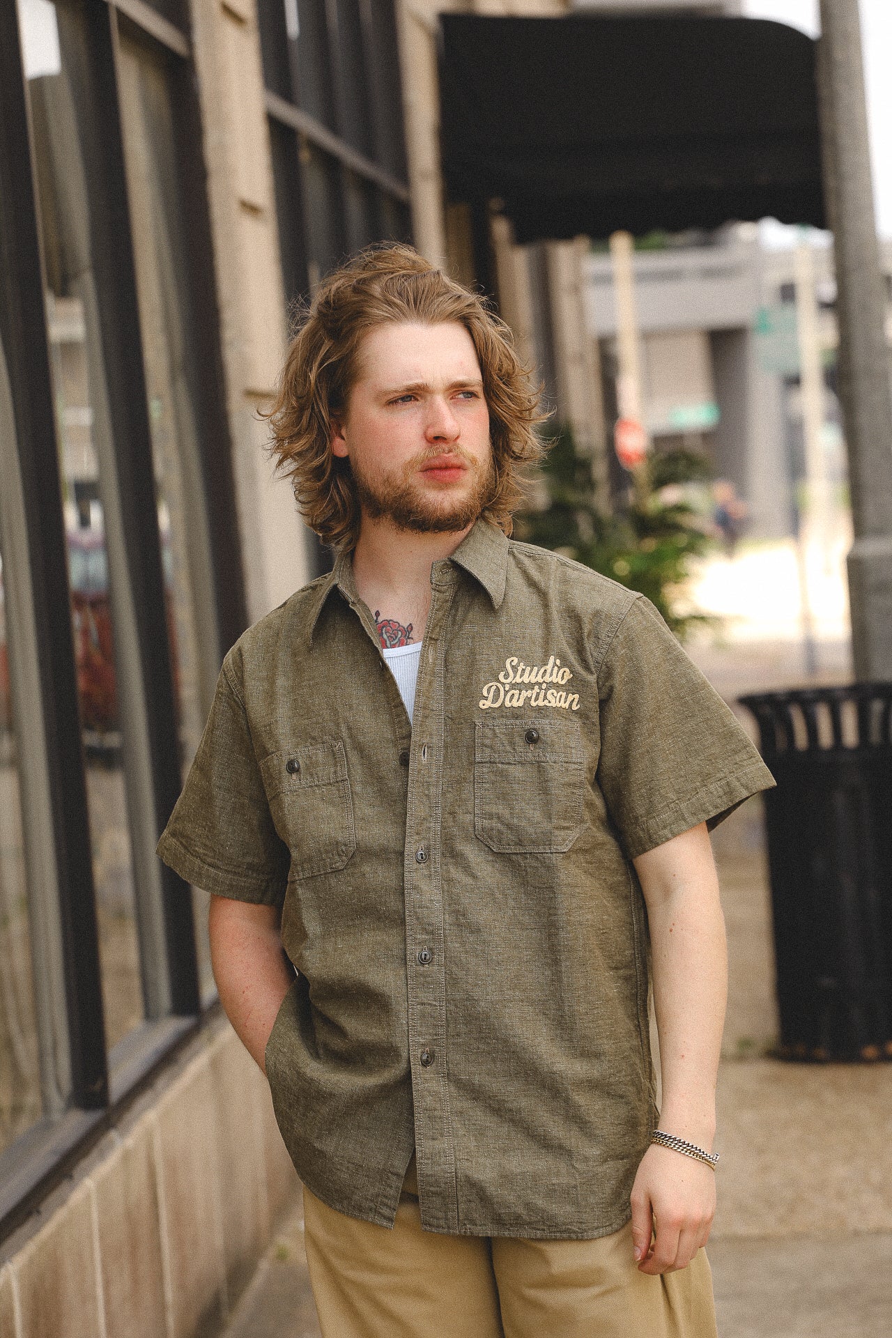 Man with beard and long hair wearing olive Studio D'artisan short sleeve shirt and beige pants on city sidewalk