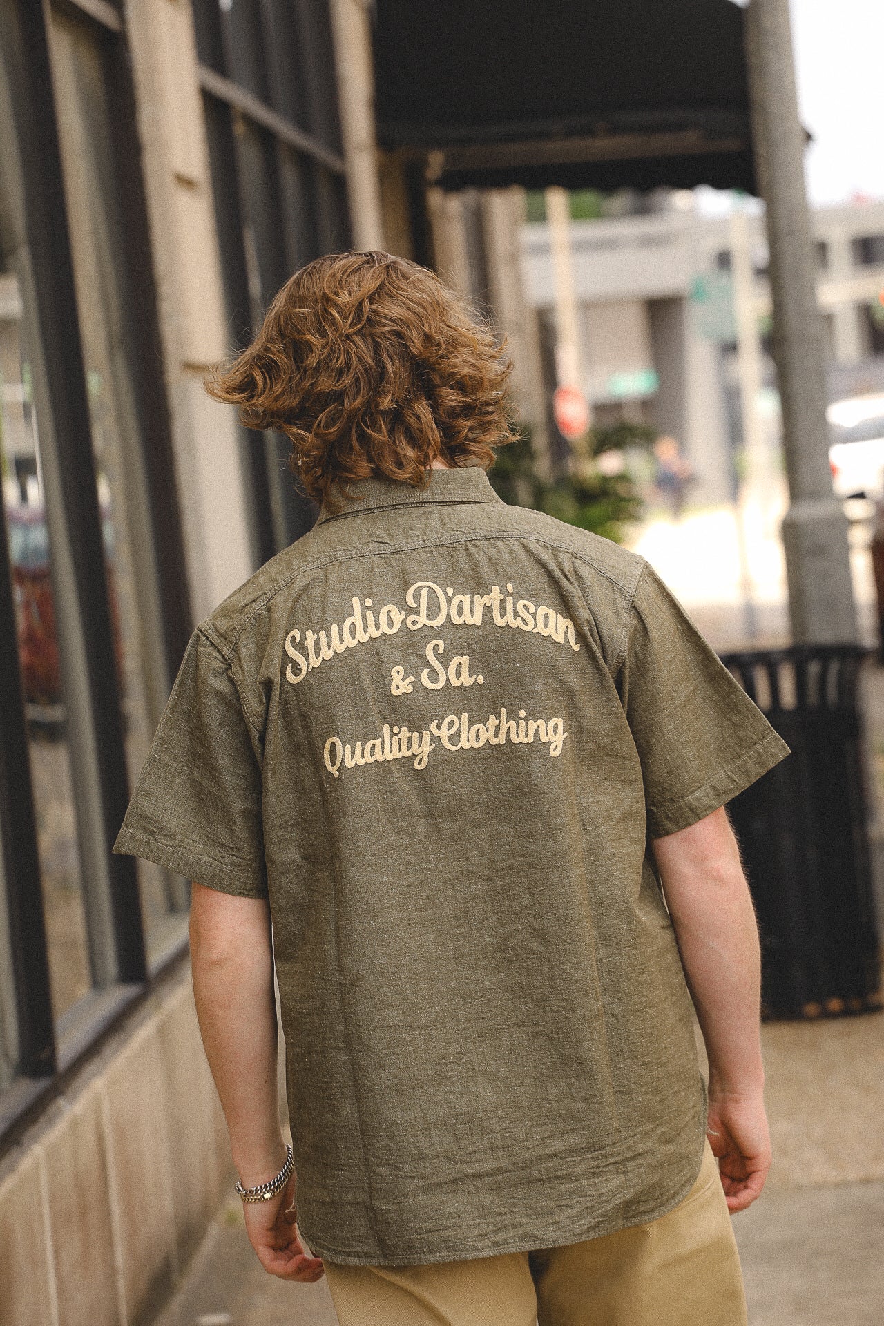Back view of person wearing olive green Studio D'artisan short sleeve shirt with beige text on city sidewalk