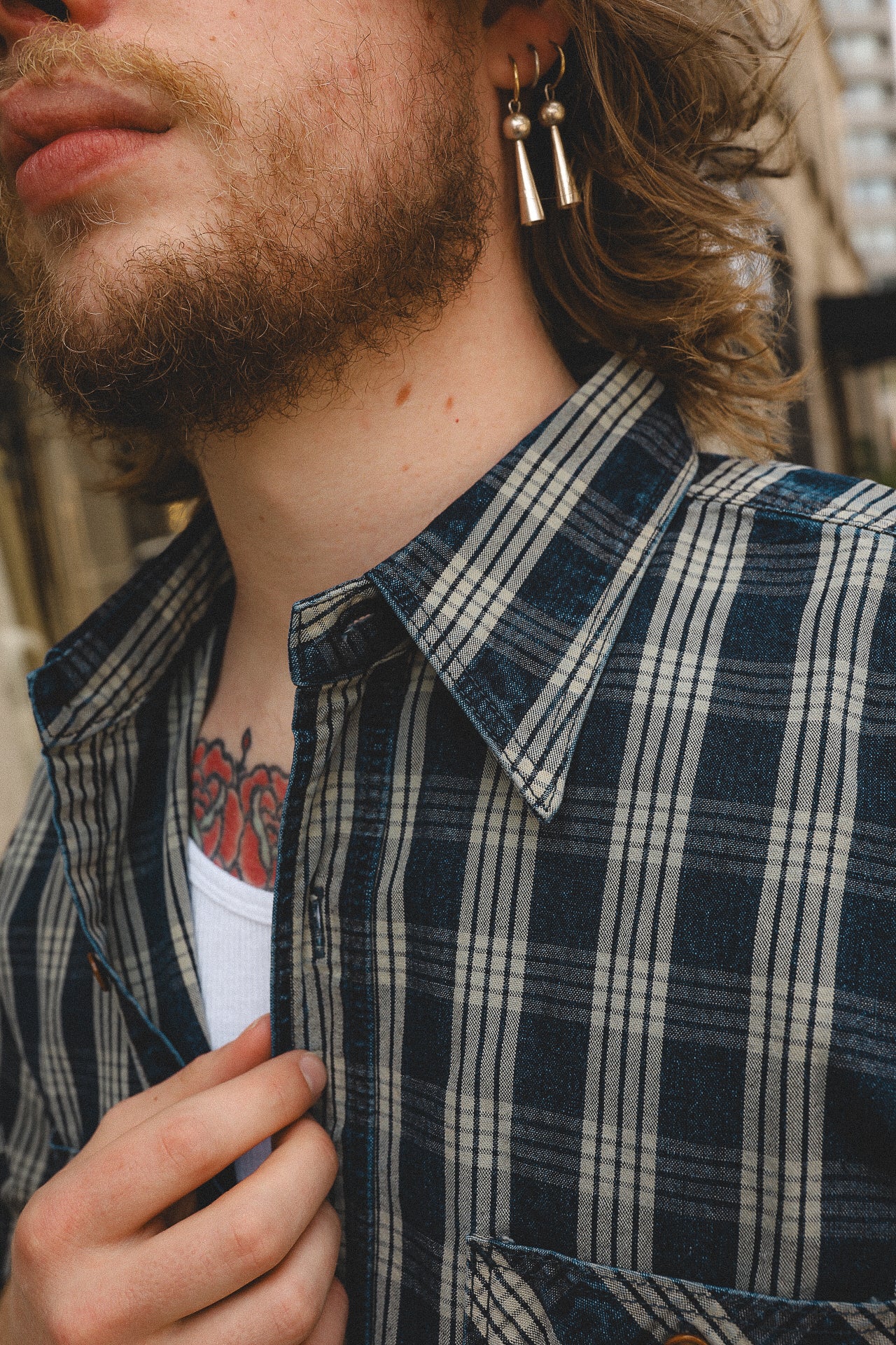 Close-up of man wearing blue plaid shirt and metal earrings, showing beard and red tattoo on chest