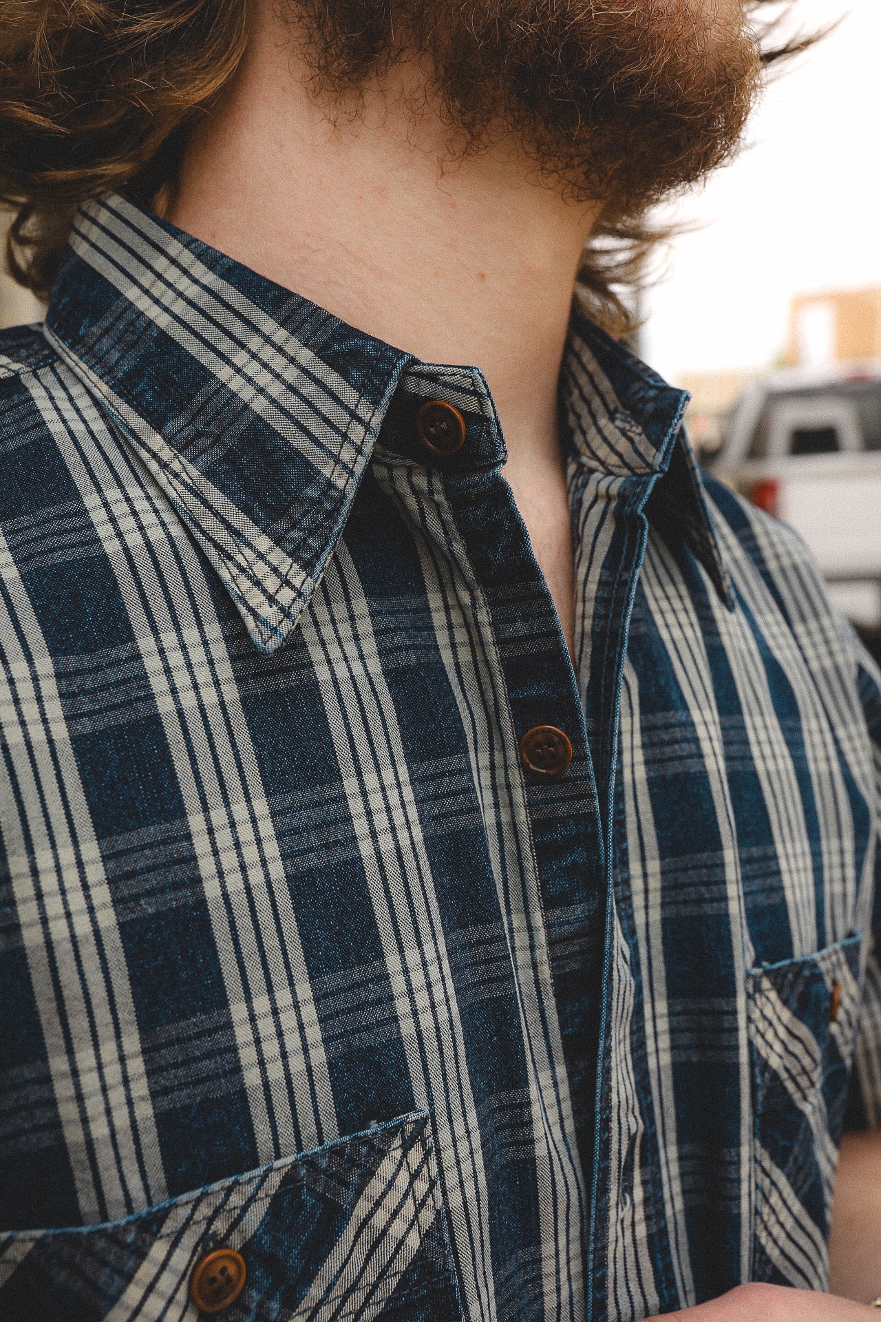 Close-up of a man wearing a blue and white plaid shirt with brown buttons and a pocket detail