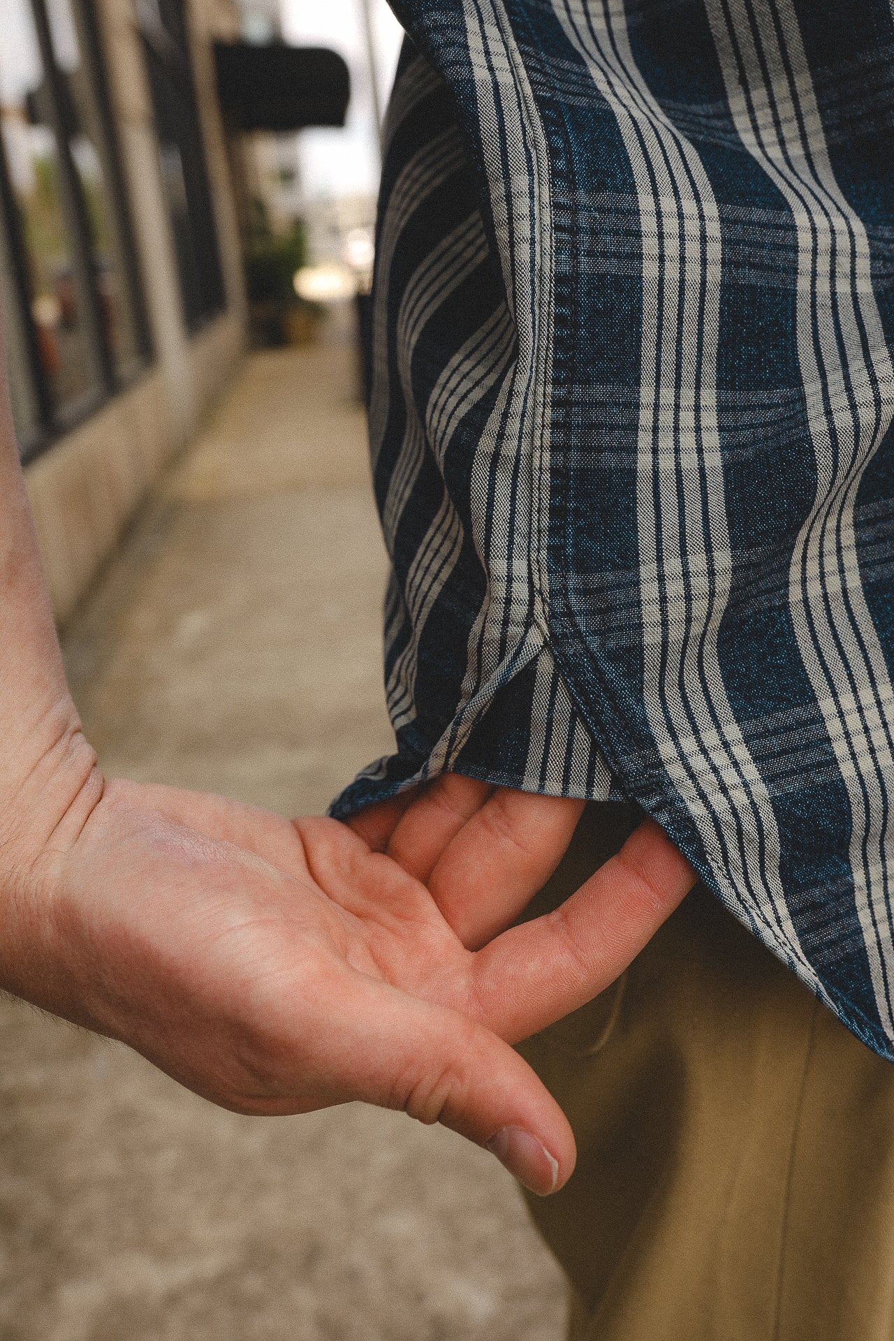 Close-up of a hand adjusting a blue and white plaid shirt near beige pants on a sidewalk