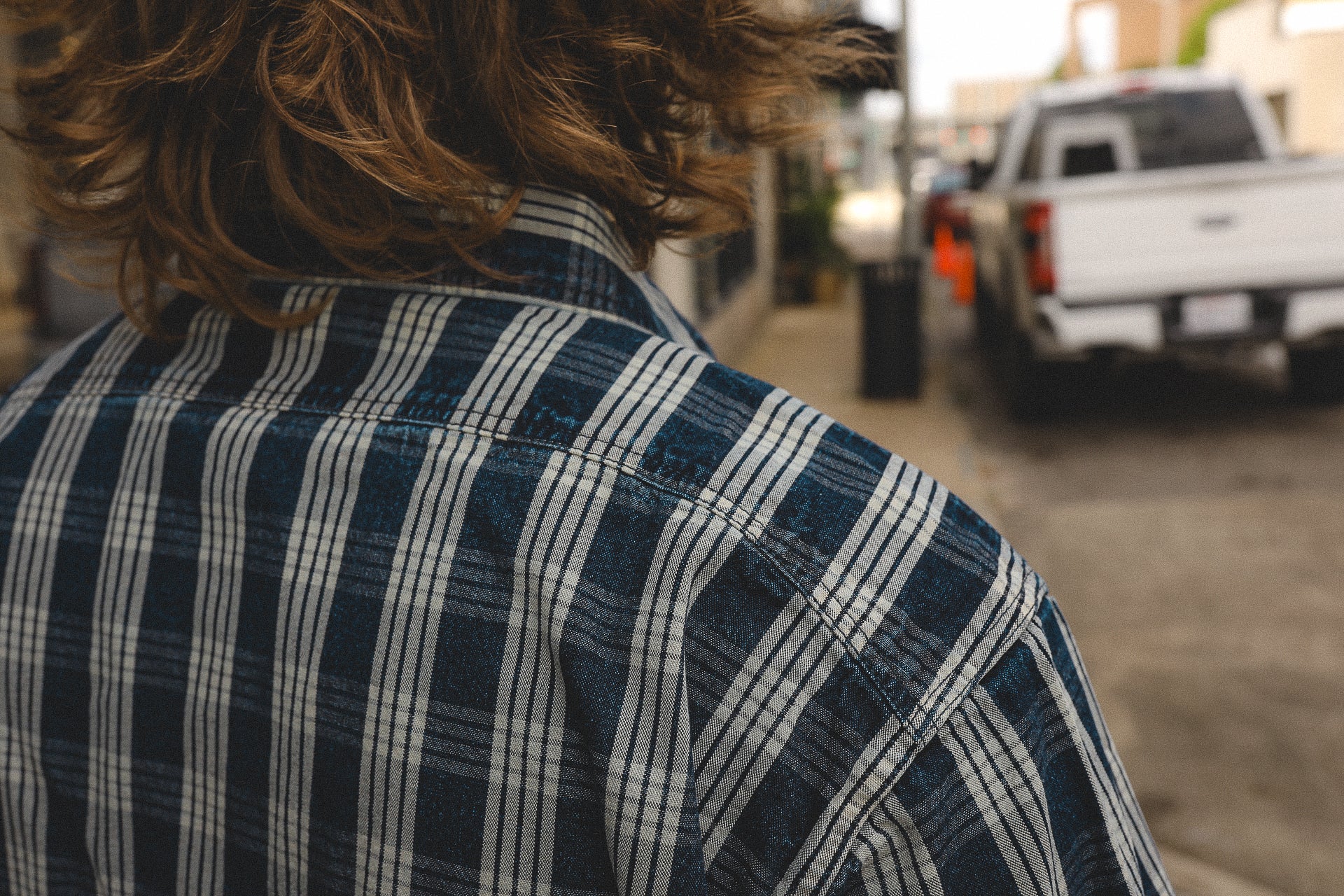 Back view of person wearing navy blue and white striped flannel shirt near a white pickup truck on street