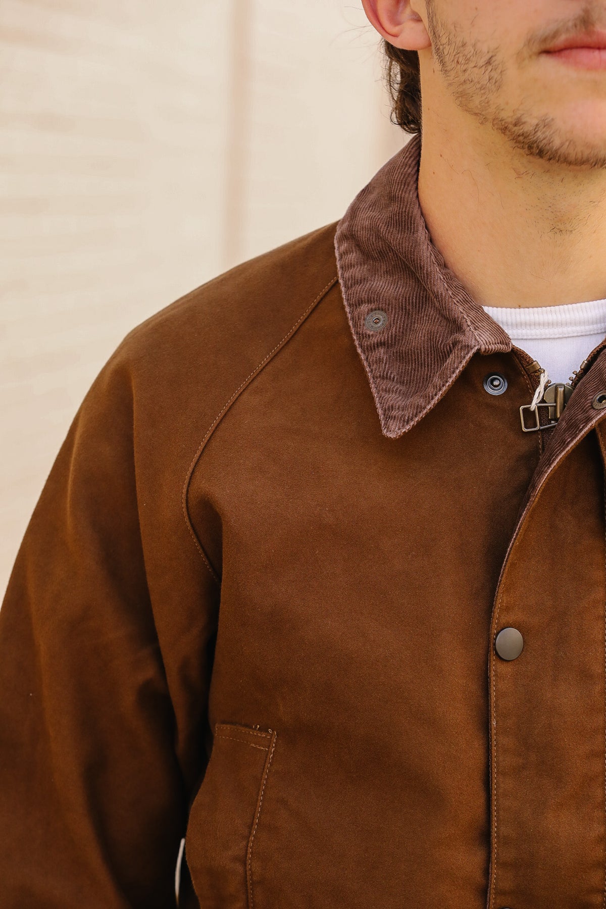 Close-up of a person wearing a brown corduroy collar overshirt or jacket over a white undershirt, with a light neutral wall in the background.