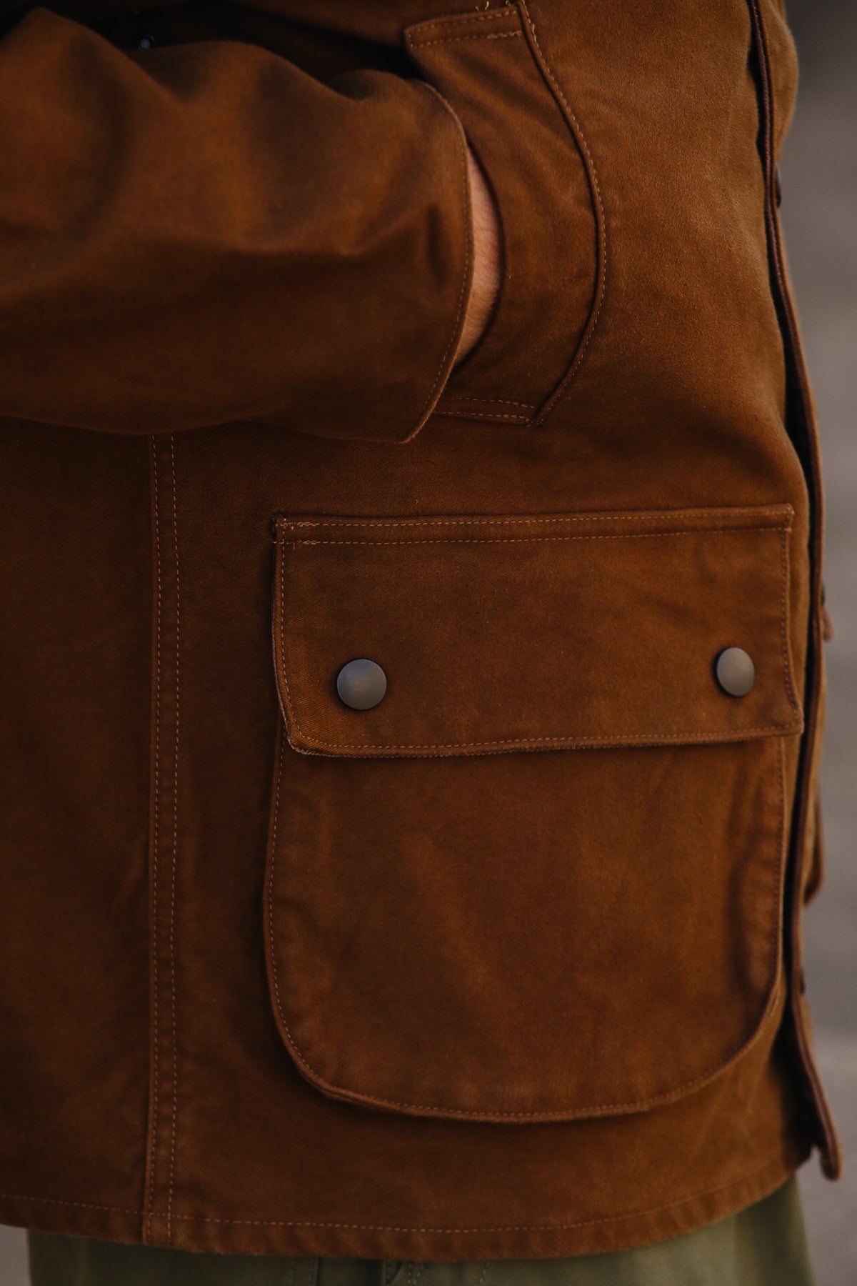 Close-up of a brown suede or suede-like jacket with a large front flap pocket secured by two metal snap buttons, worn by a person.