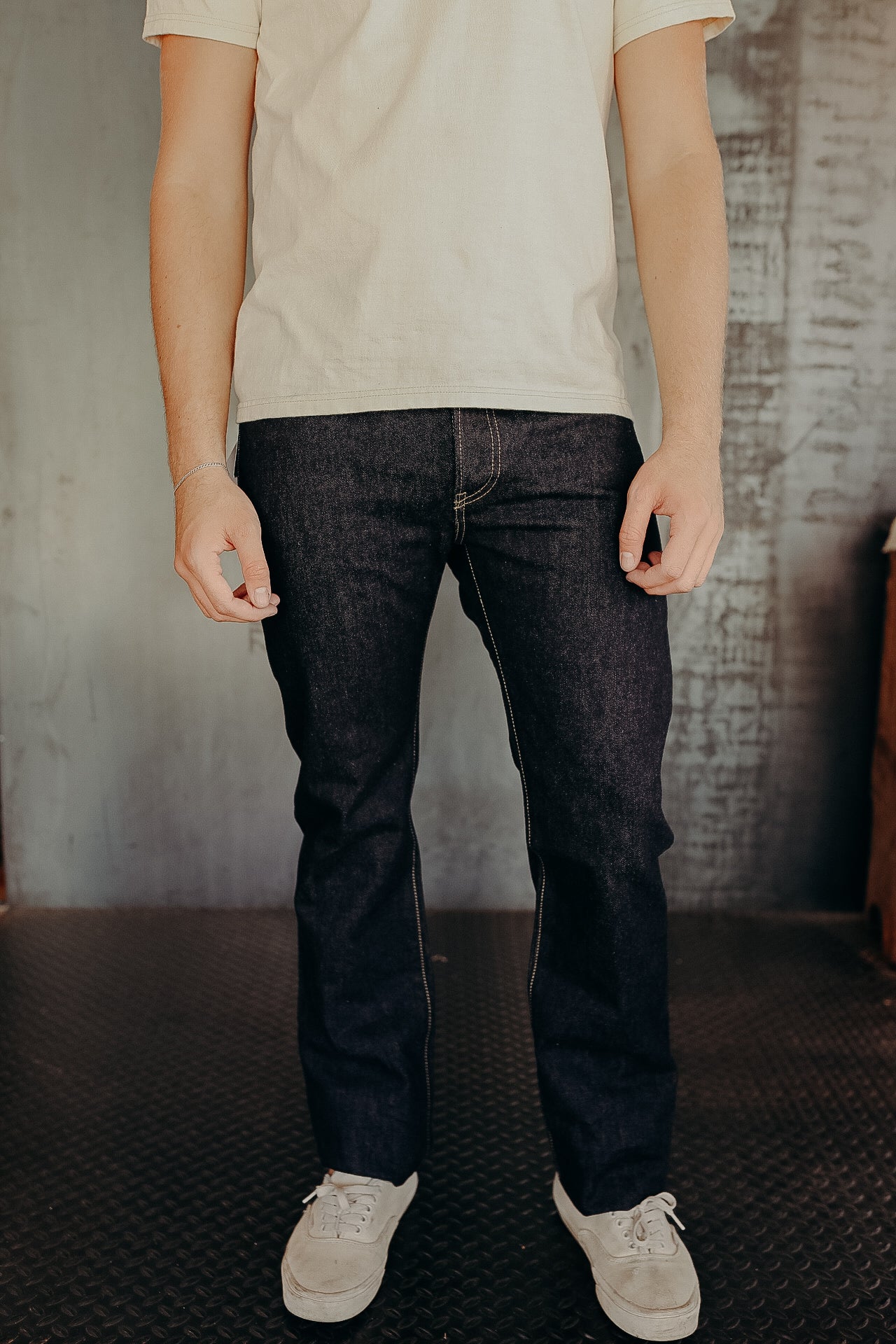 Man wearing raw denim jeans with white sneakers and a beige t-shirt in industrial setting