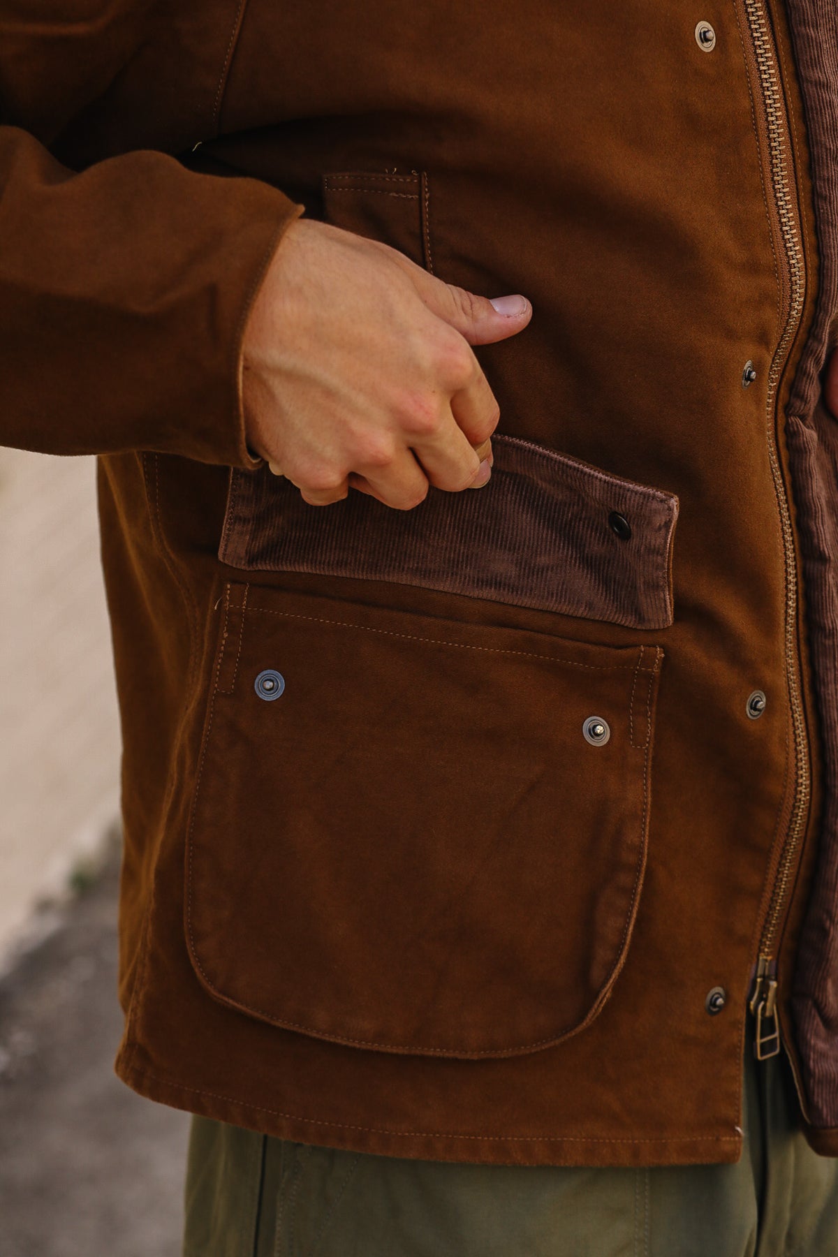 Close-up of a person wearing a brown waxed canvas jacket, showing a hand reaching into a front utility pocket with a corduroy flap.