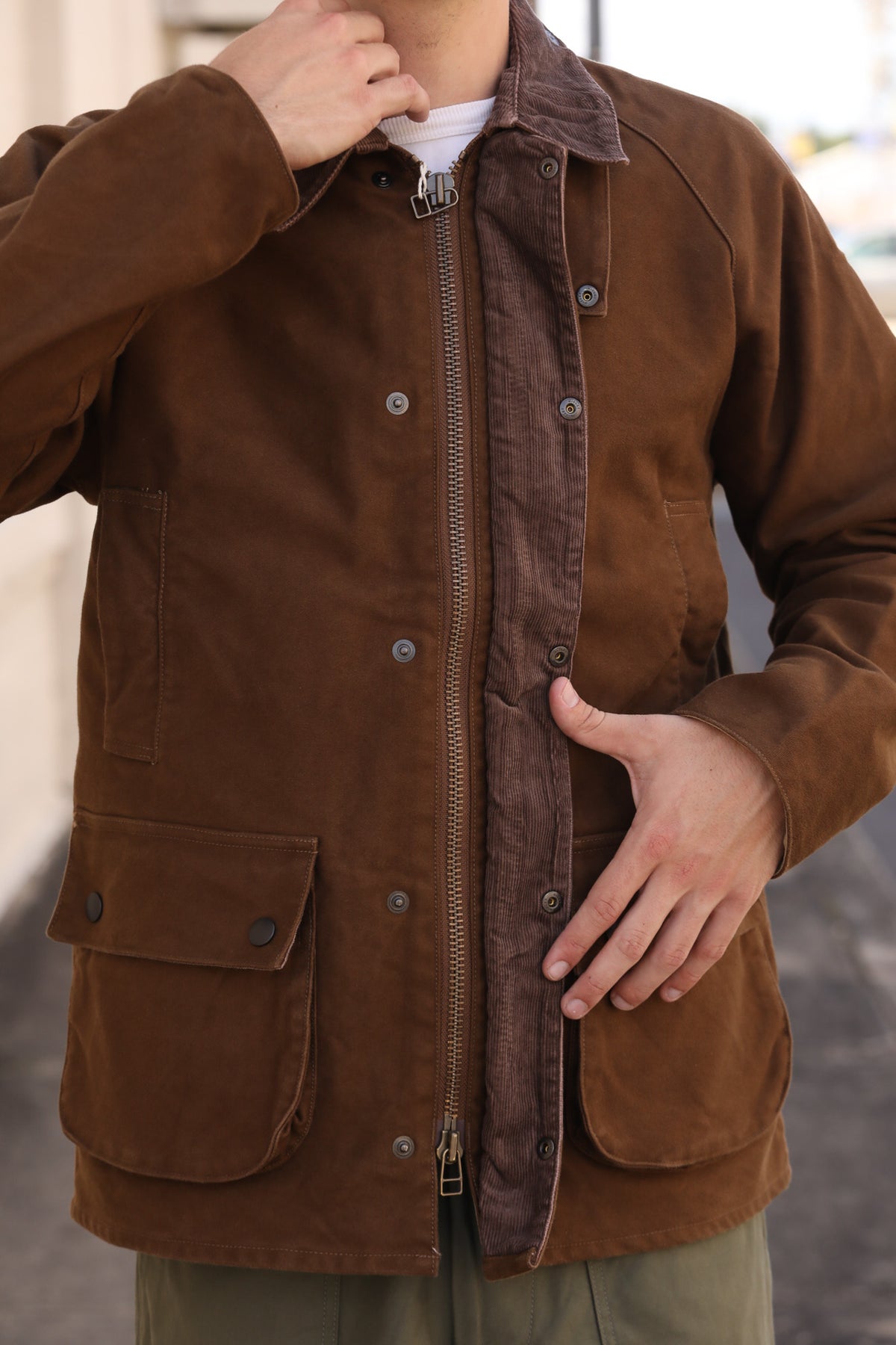A man wearing a brown waxed canvas utility jacket with a front zipper and button snap closure, layered over a white t-shirt and olive green pants.