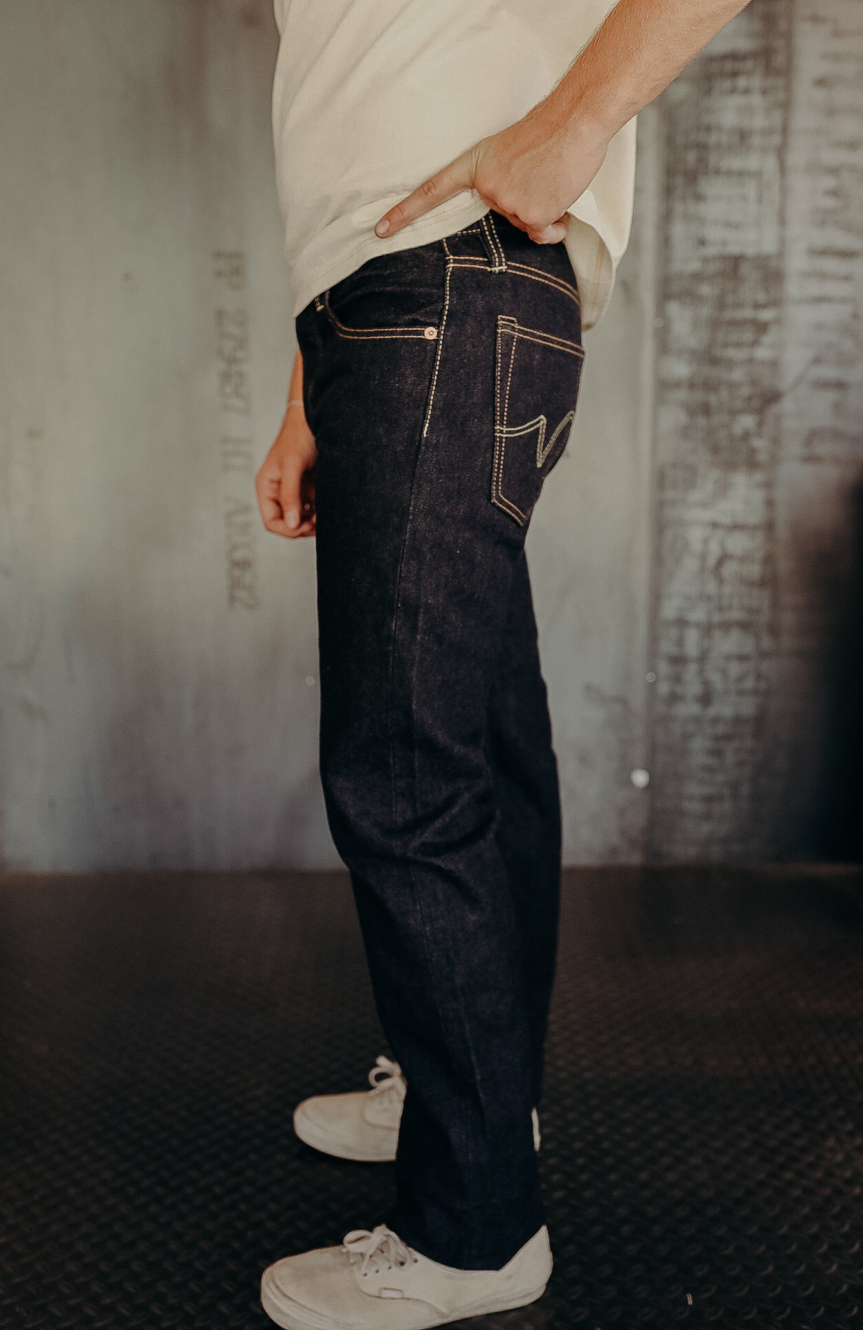 Side view of person wearing dark denim jeans with contrast stitching and white sneakers in industrial setting