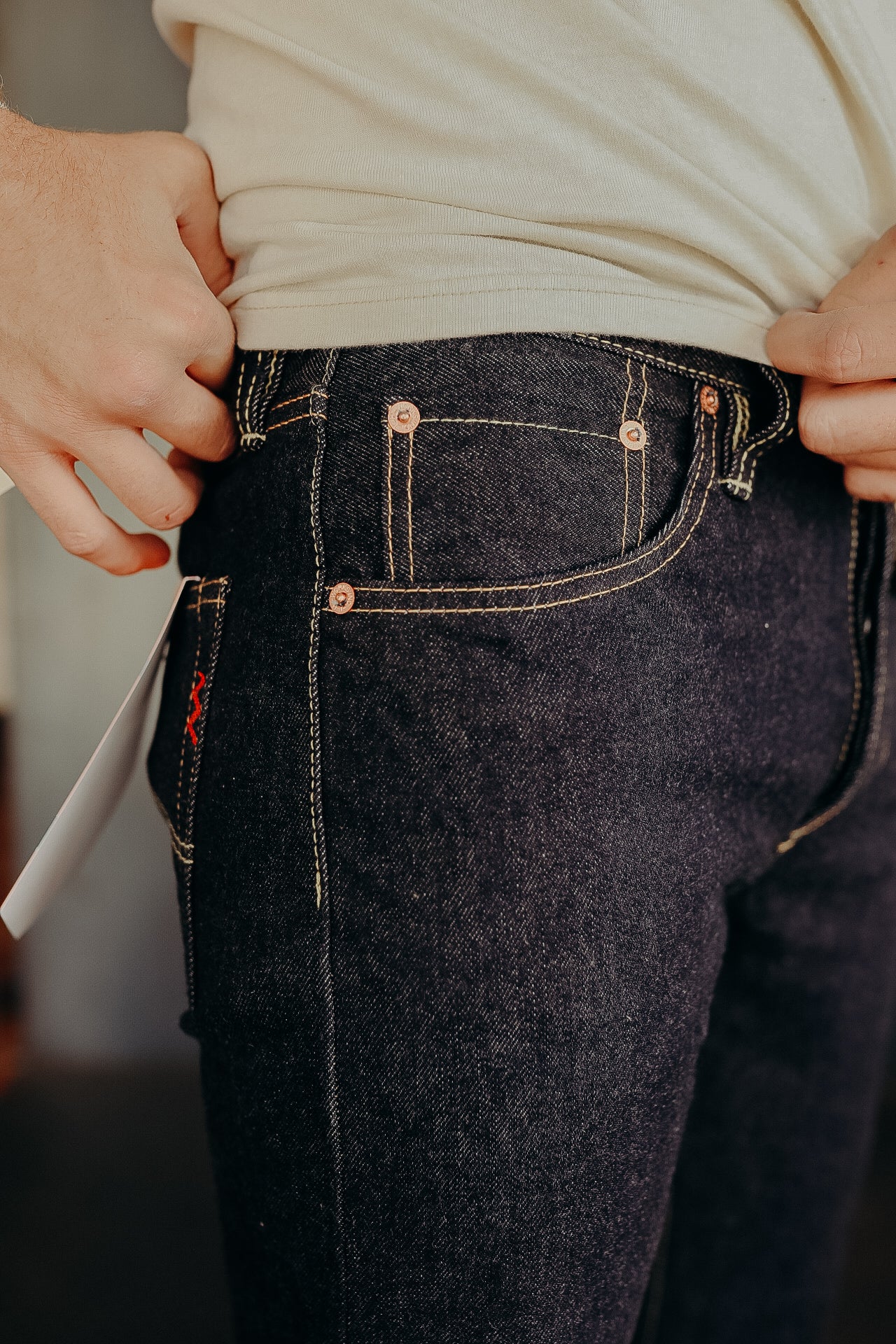 Close-up of man wearing dark raw denim jeans with copper rivets and contrast stitching, light beige shirt