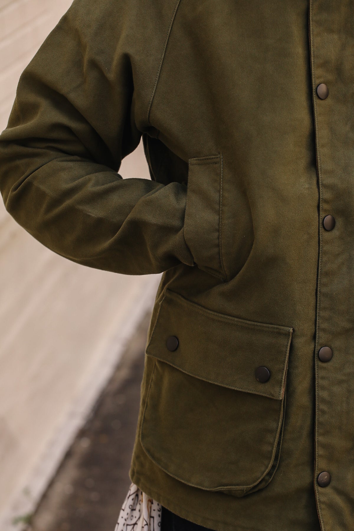Close-up of an olive green waxed canvas utility jacket with dark metal snap buttons and a front flap pocket, worn by a model.