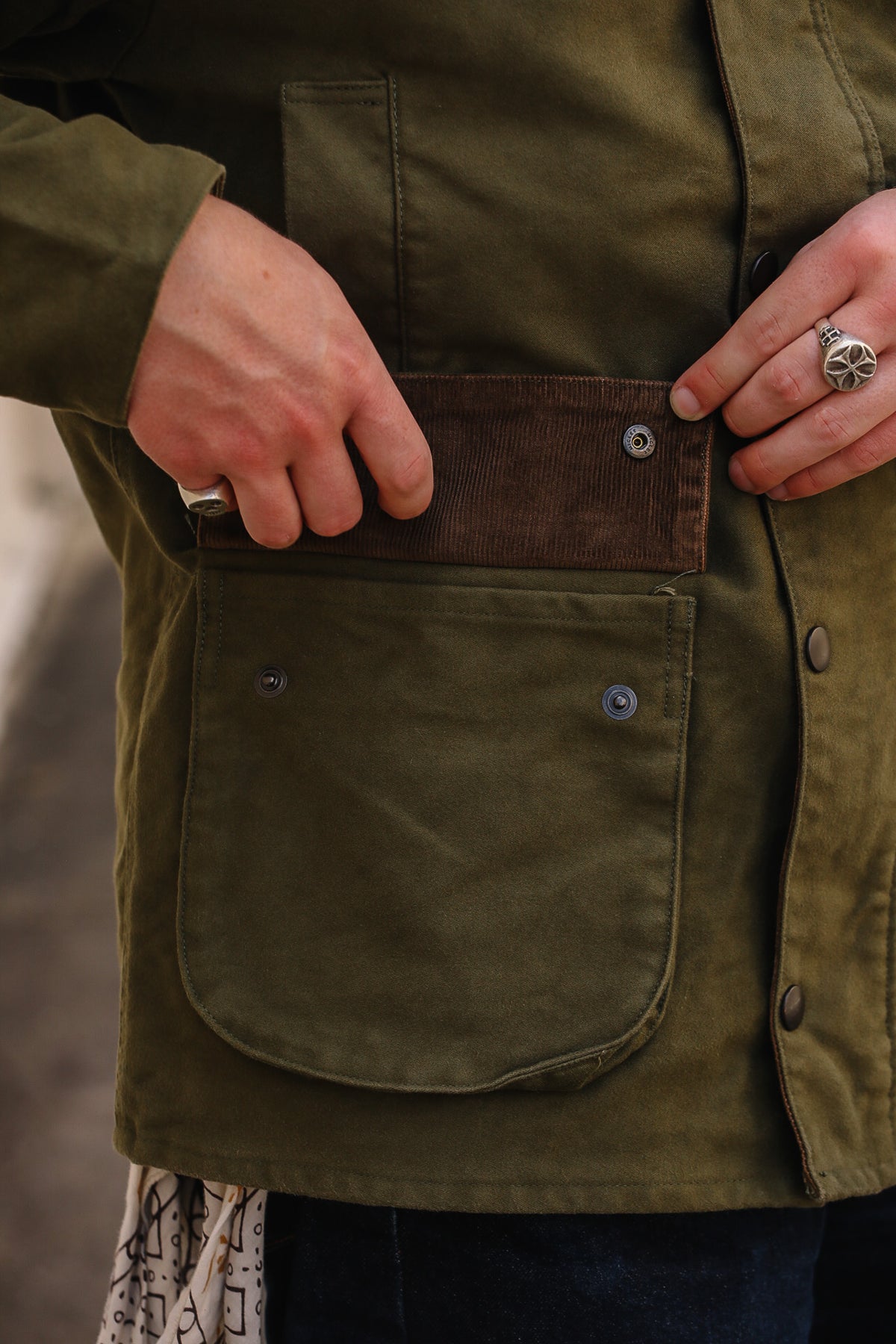 Close-up of a person wearing an olive green waxed canvas jacket, showing a front utility pocket with a brown corduroy flap and metal snap buttons.