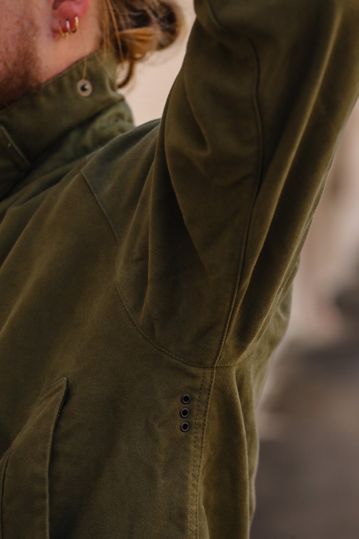 Close-up of a person wearing an olive green jacket, showing the sleeve, shoulder, and chest area with a blurred outdoor background.