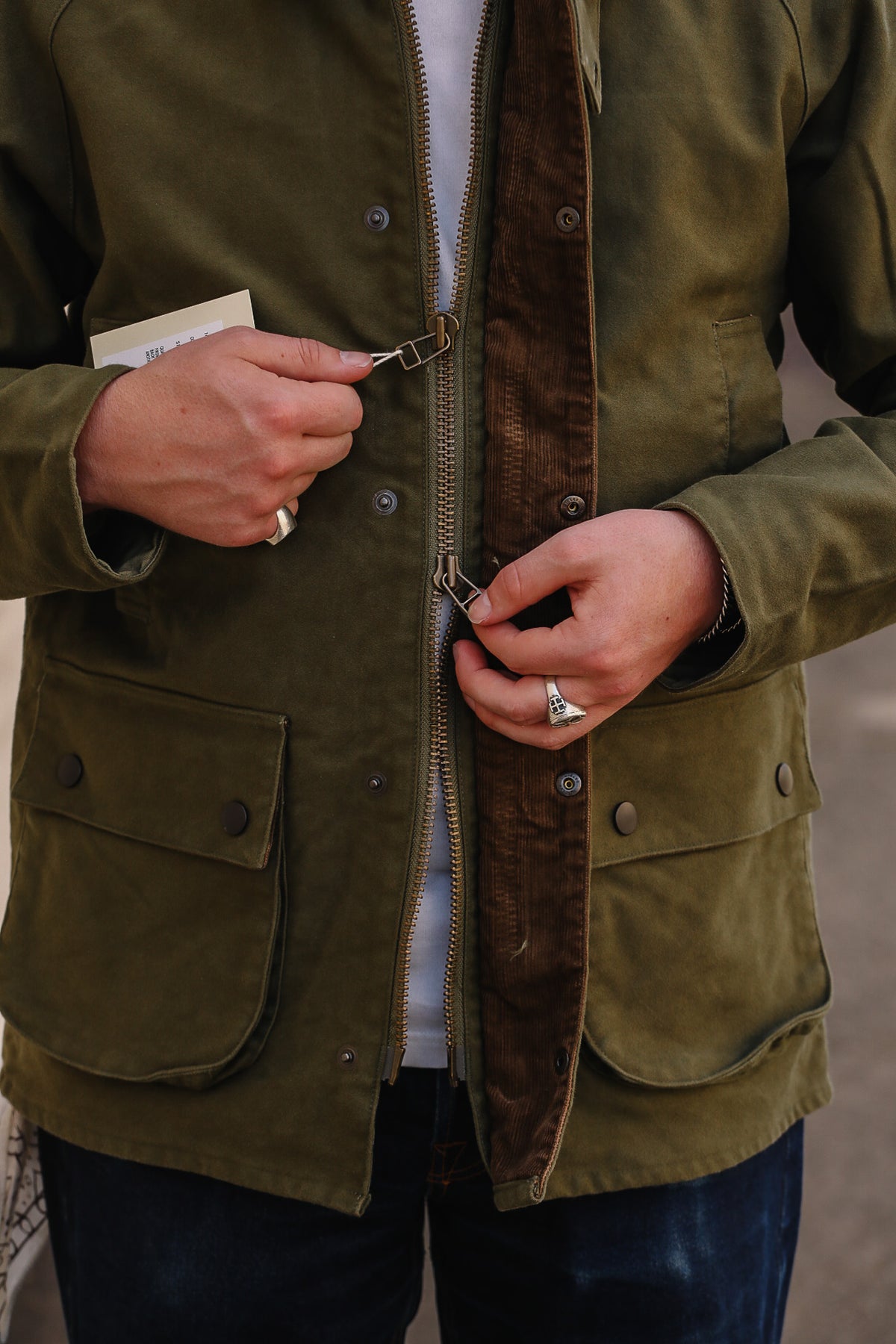 Close-up of a person wearing an olive green waxed canvas utility jacket with a brown corduroy-lined front, zipped and buttoned, paired with a white shirt and dark blue jeans.