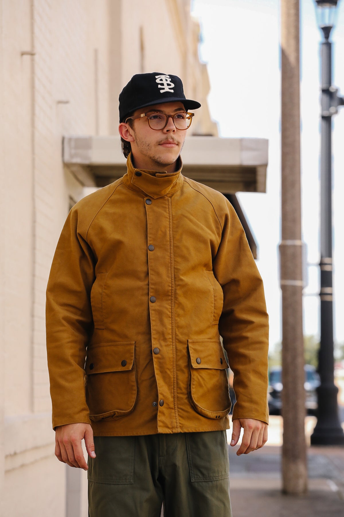 A man wearing a mustard brown waxed canvas utility jacket, olive green pants, a black baseball cap, and glasses, standing outdoors on a city street.