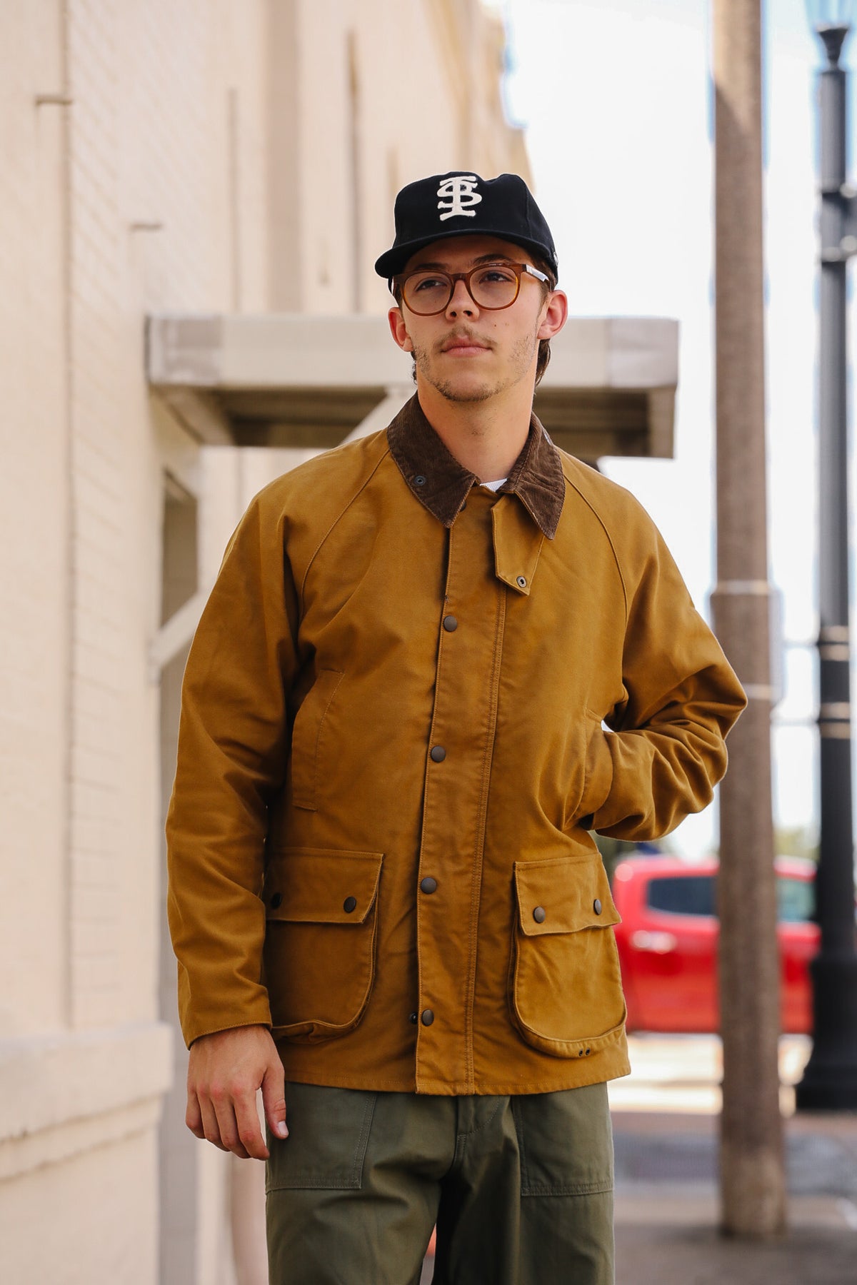 A man wearing a mustard yellow waxed cotton utility jacket with a brown corduroy collar, olive green pants, and a black baseball cap with a white logo, standing outdoors on a city street.