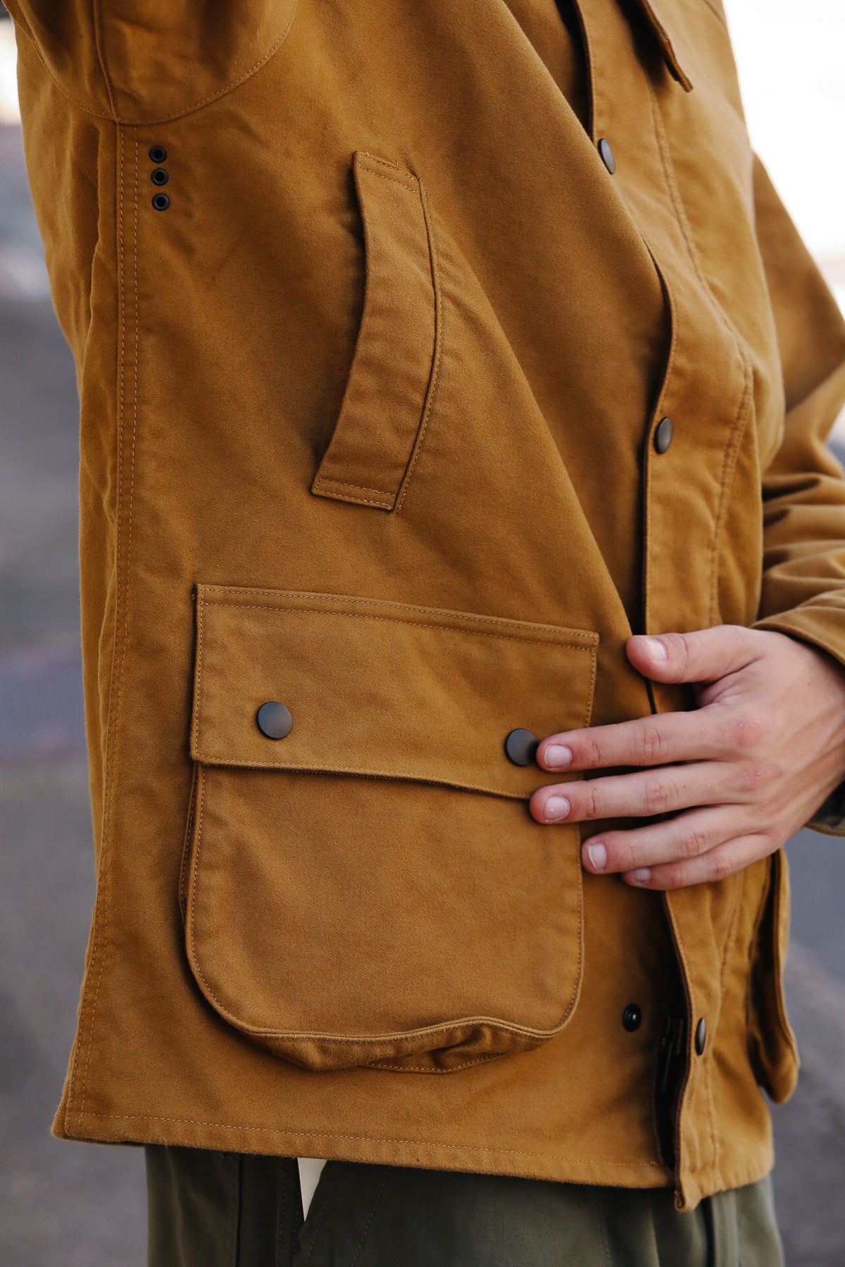 Close-up of a person wearing a mustard brown waxed canvas utility jacket, showing a front flap pocket with snap buttons and a hand resting on the pocket.