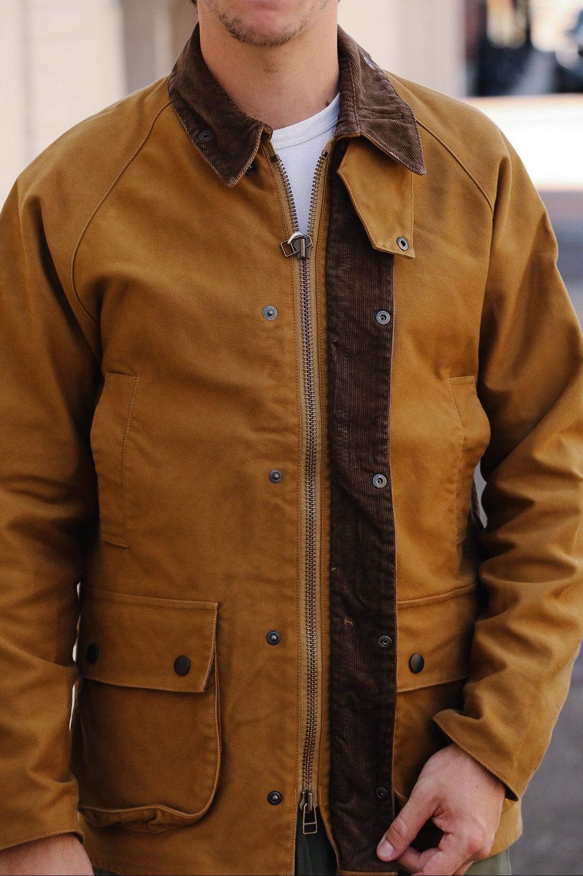 A man wearing a brown waxed canvas utility jacket with a dark brown corduroy collar and a front zipper, layered over a white t-shirt.