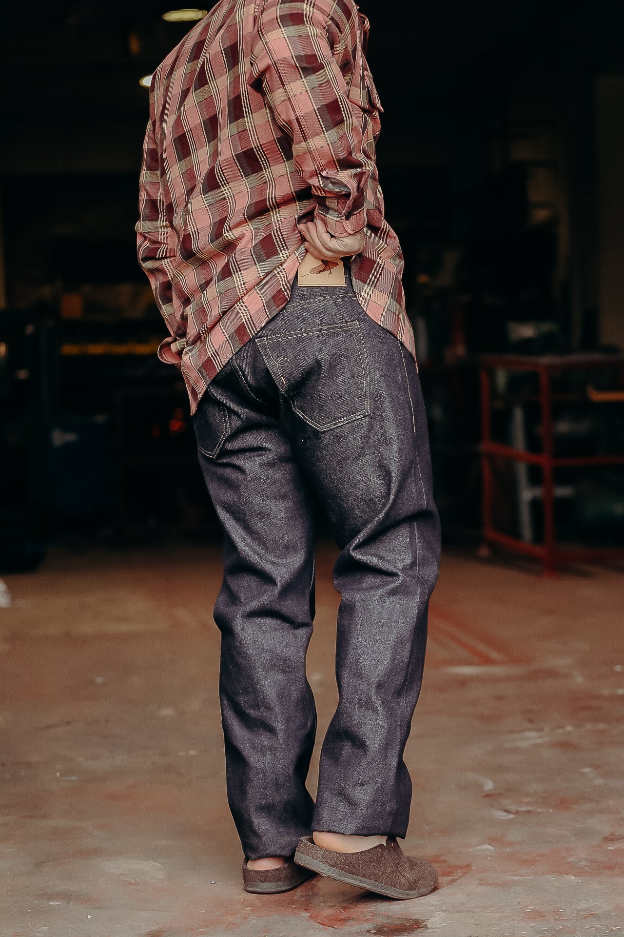 Person wearing dark raw denim jeans and a red plaid shirt in a workshop setting