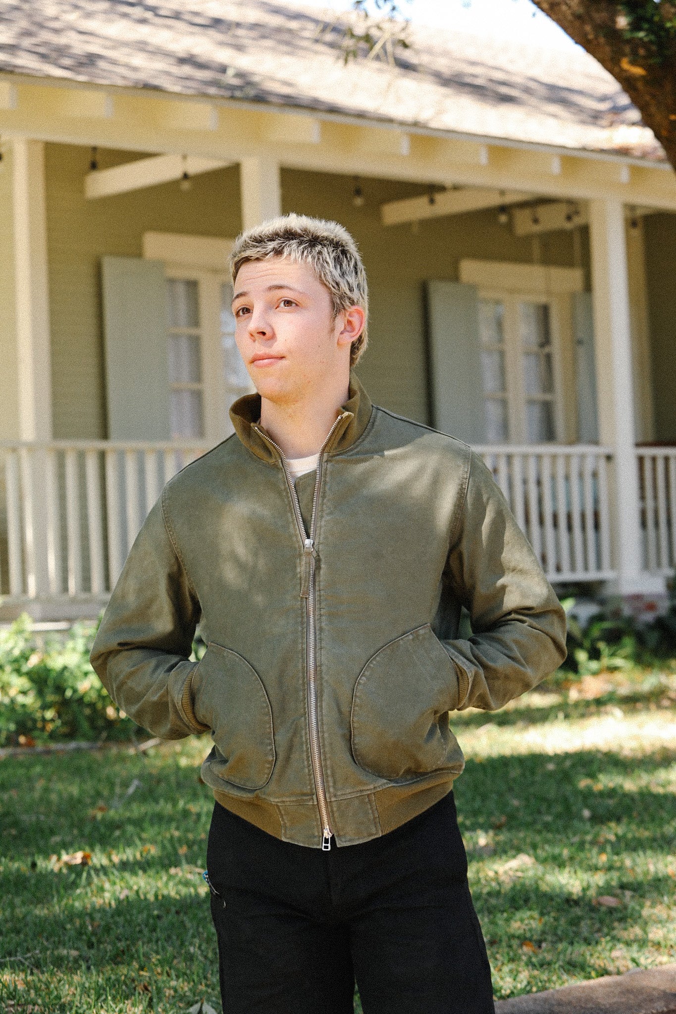 Young man wearing olive green zip-up jacket standing outside a house with porch and greenery