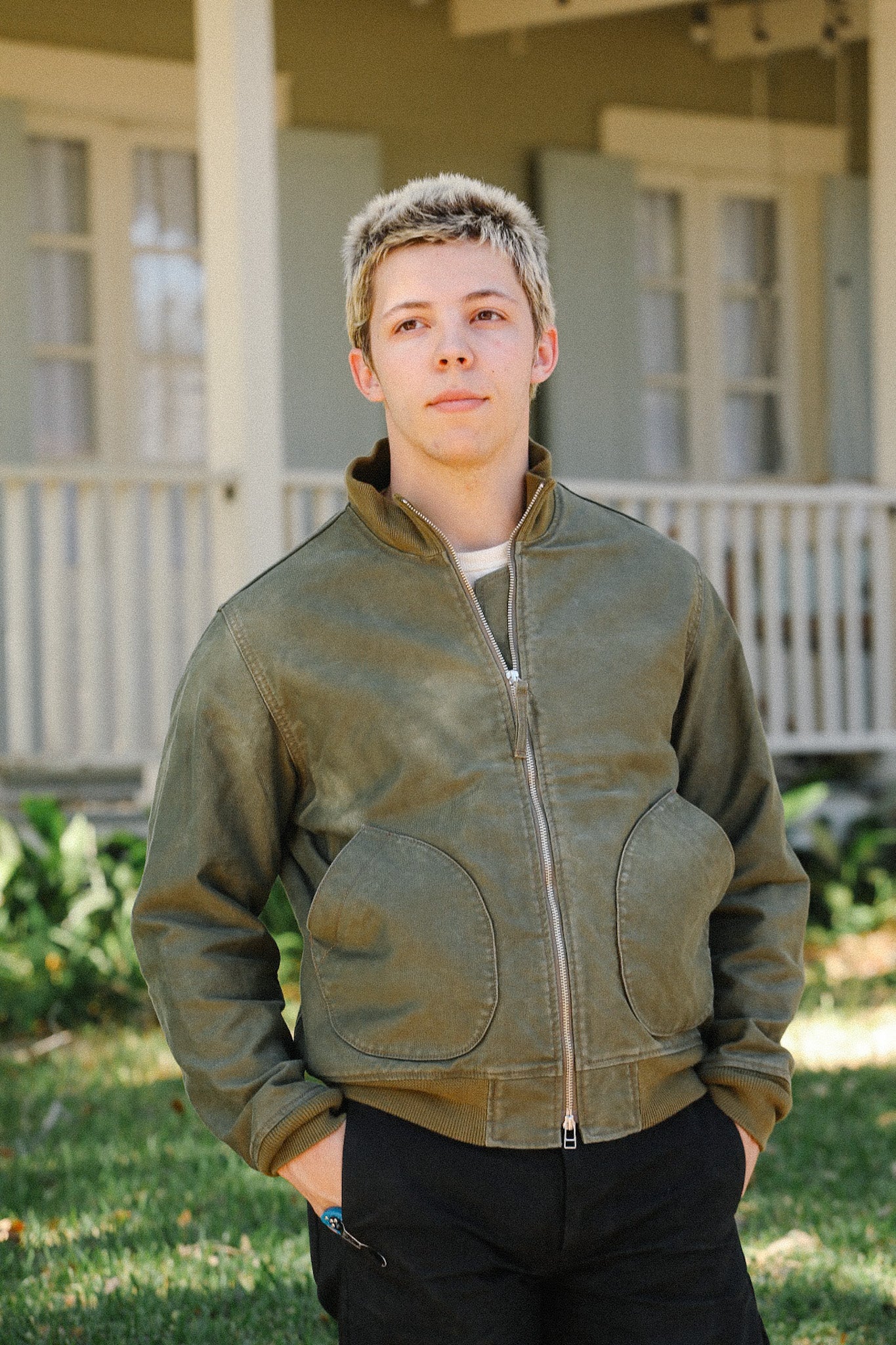 Young man wearing green zip-up jacket with large front pockets standing outdoors near a porch