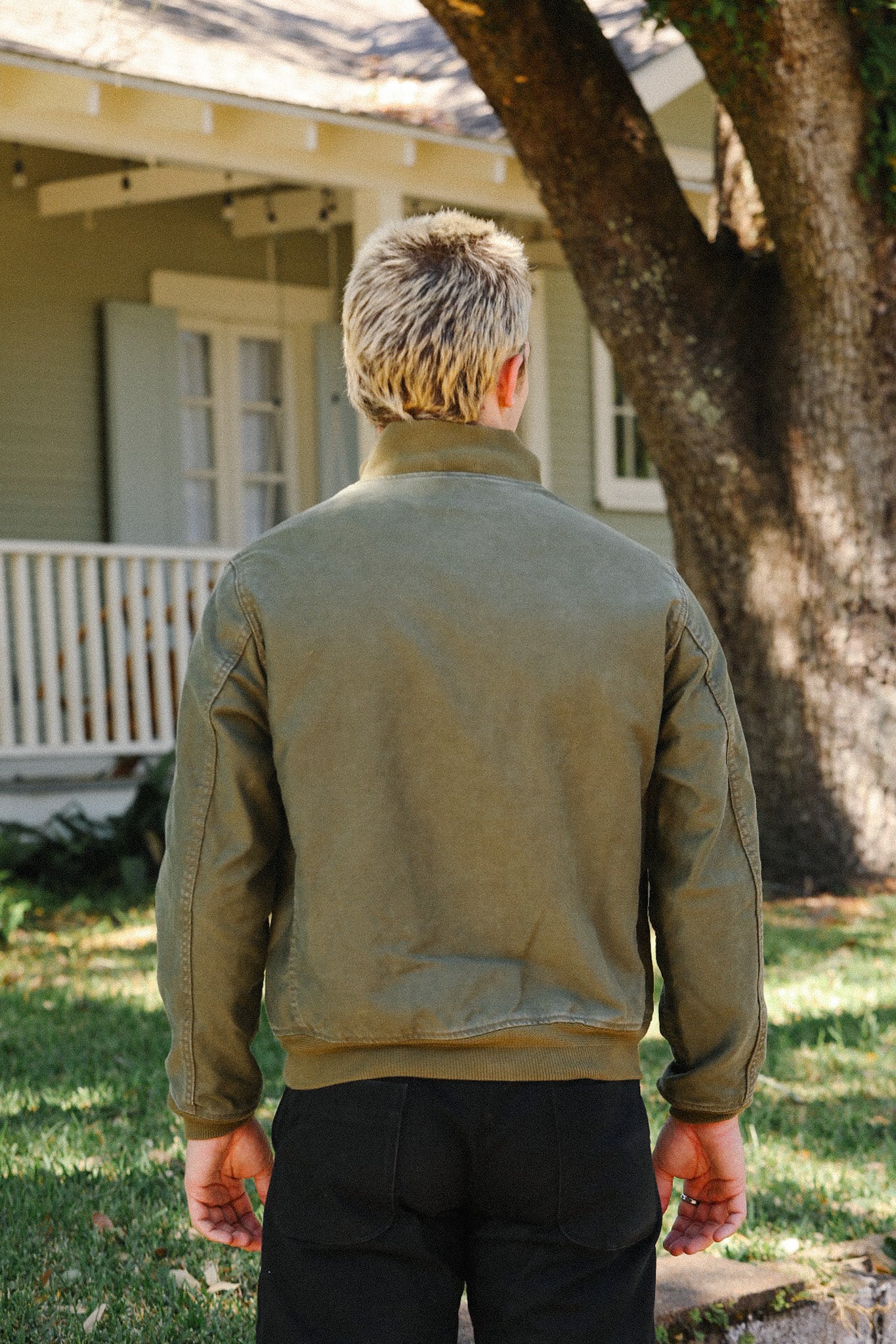 Man with short blond hair wearing a green jacket and black pants outdoors near a tree and house