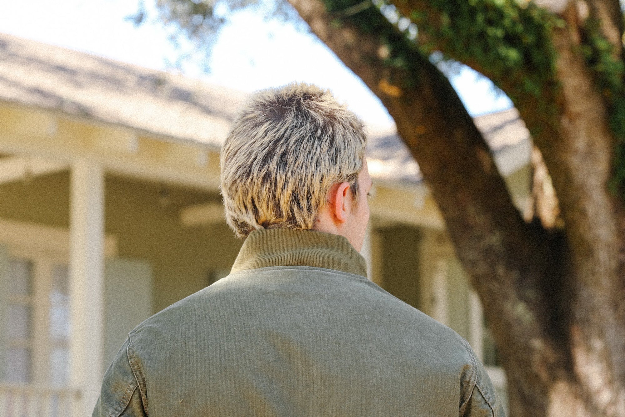 Man with short blonde hair wearing green jacket outdoors near tree and house