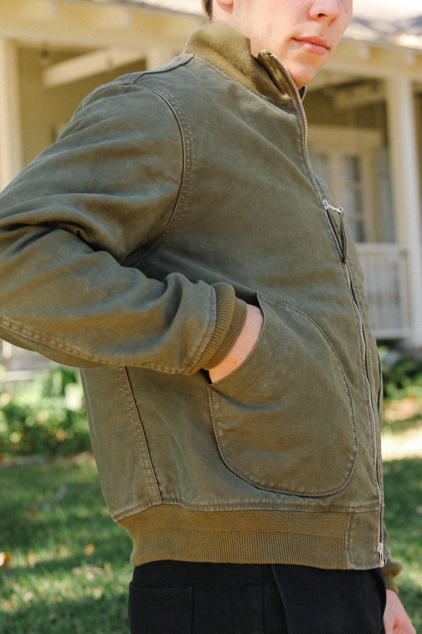 Person wearing an olive green casual jacket with zip front and pockets outside a porch house