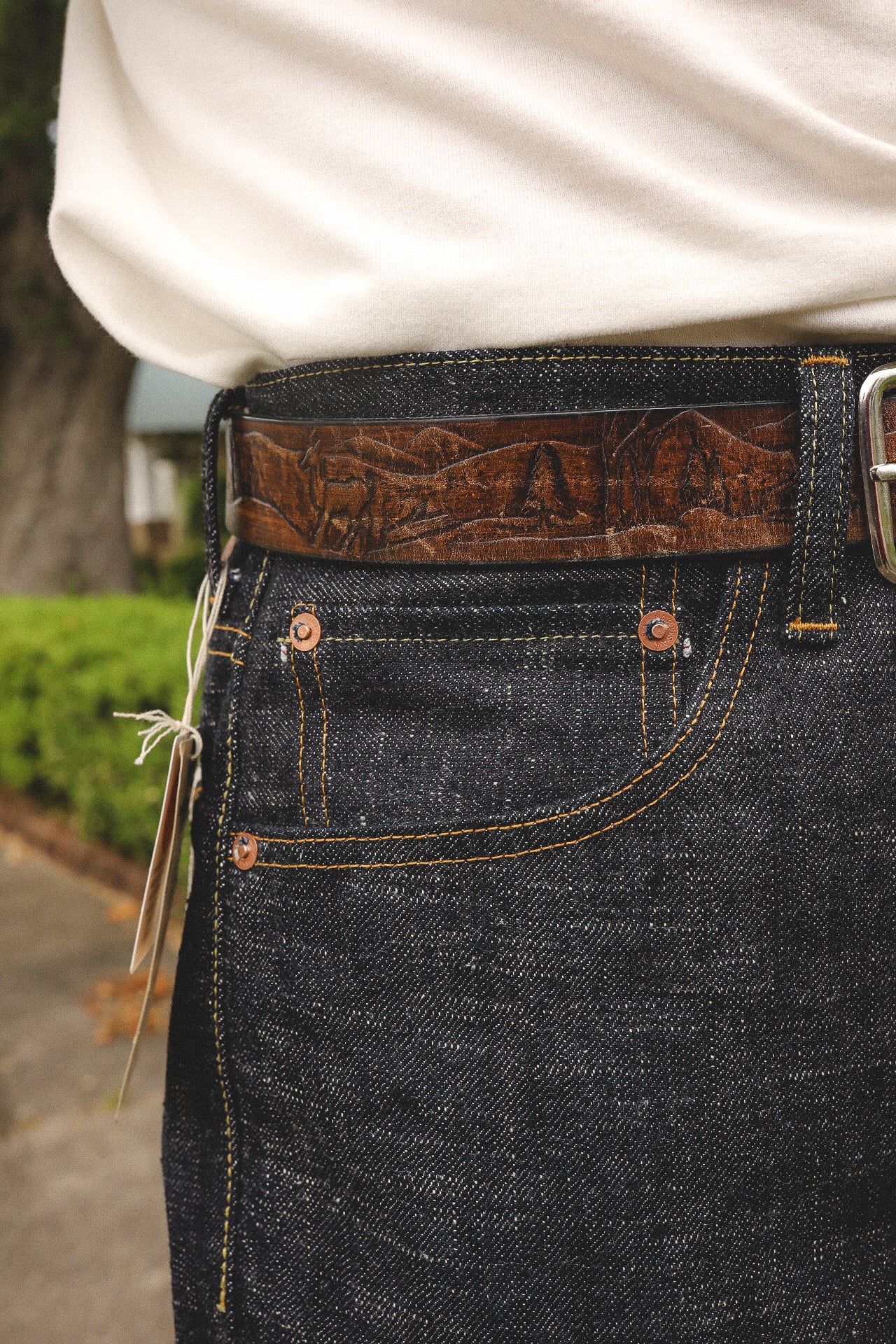 close-up of dark denim jeans with orange stitching and a brown leather belt featuring mountain and deer engraving