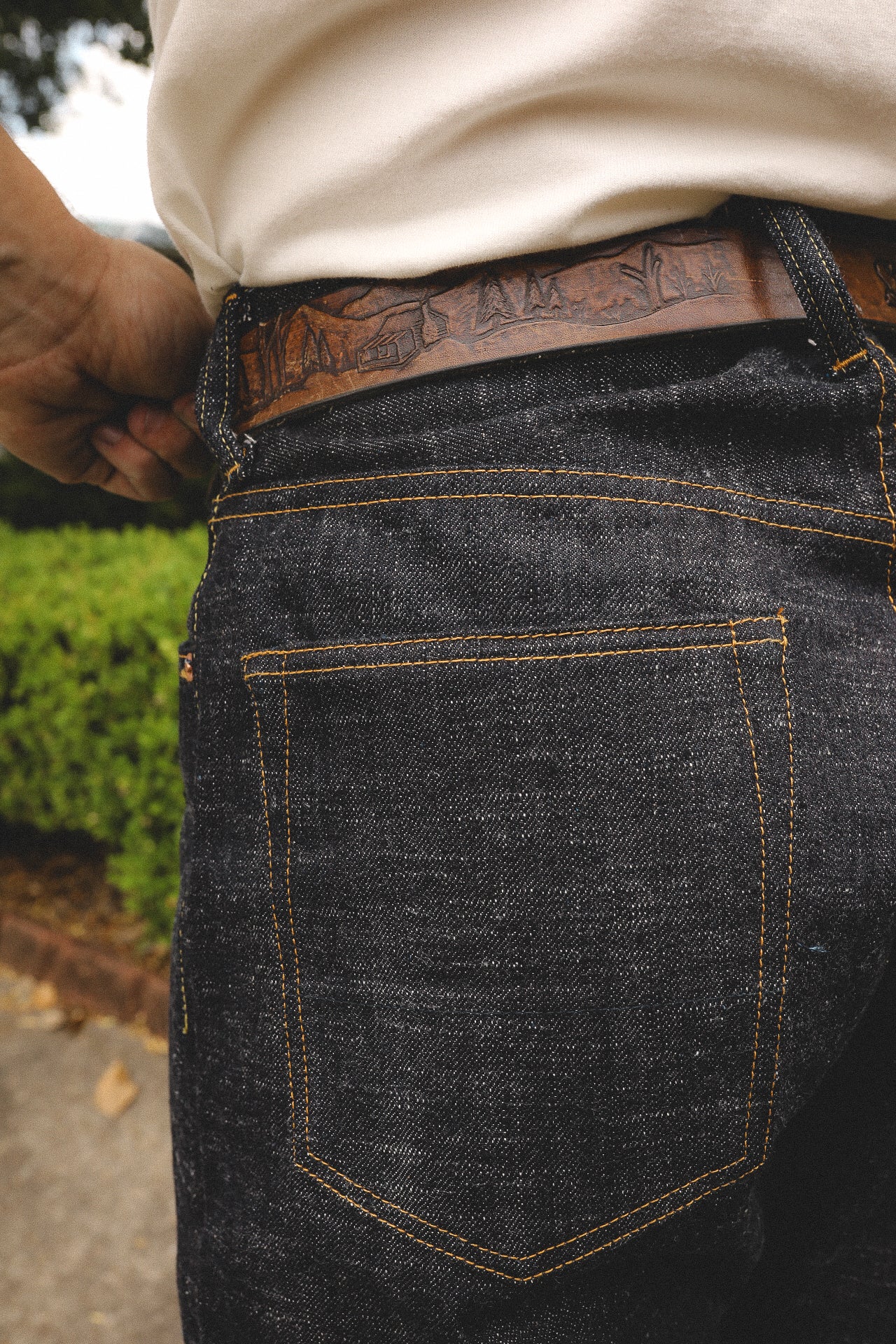 Close-up of dark denim jeans back pocket with orange stitching and rustic leather belt featuring engraved mountain scene