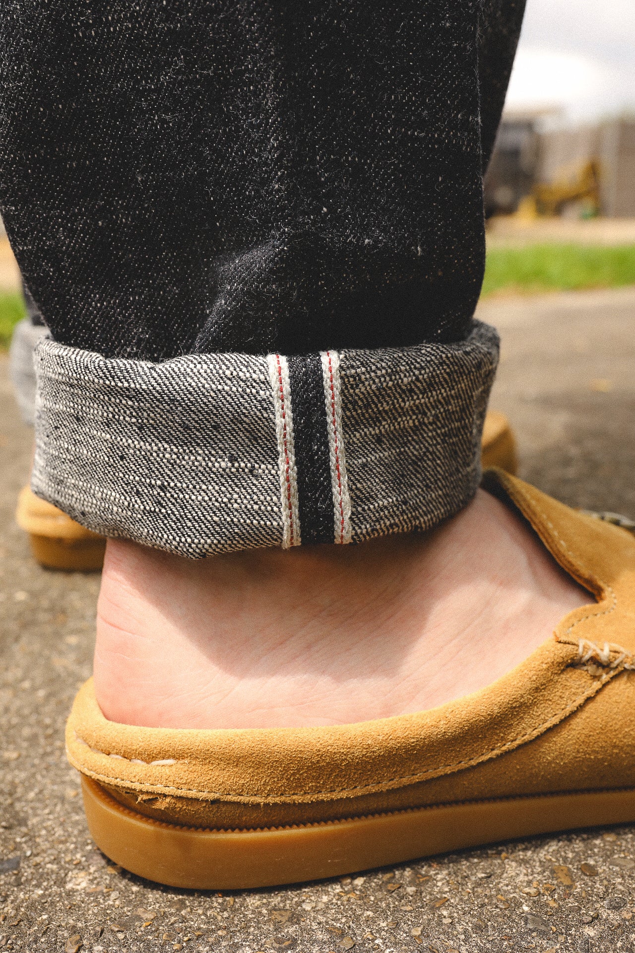 close-up of cuffed black denim jeans with selvedge edge paired with tan suede loafers on concrete surface