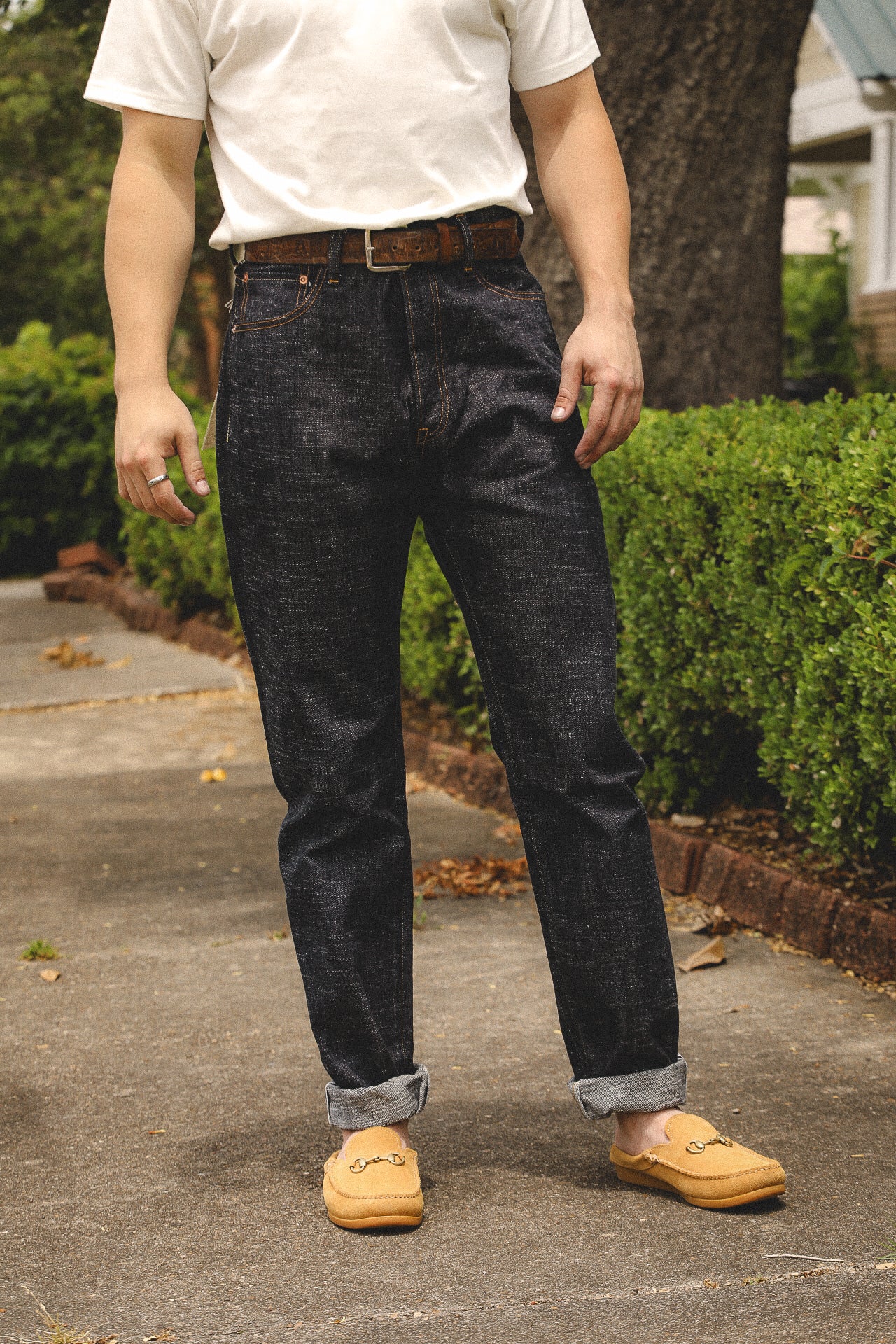 Man wearing dark denim jeans with rolled cuffs, white t-shirt, brown belt, and tan moccasin loafers outdoors