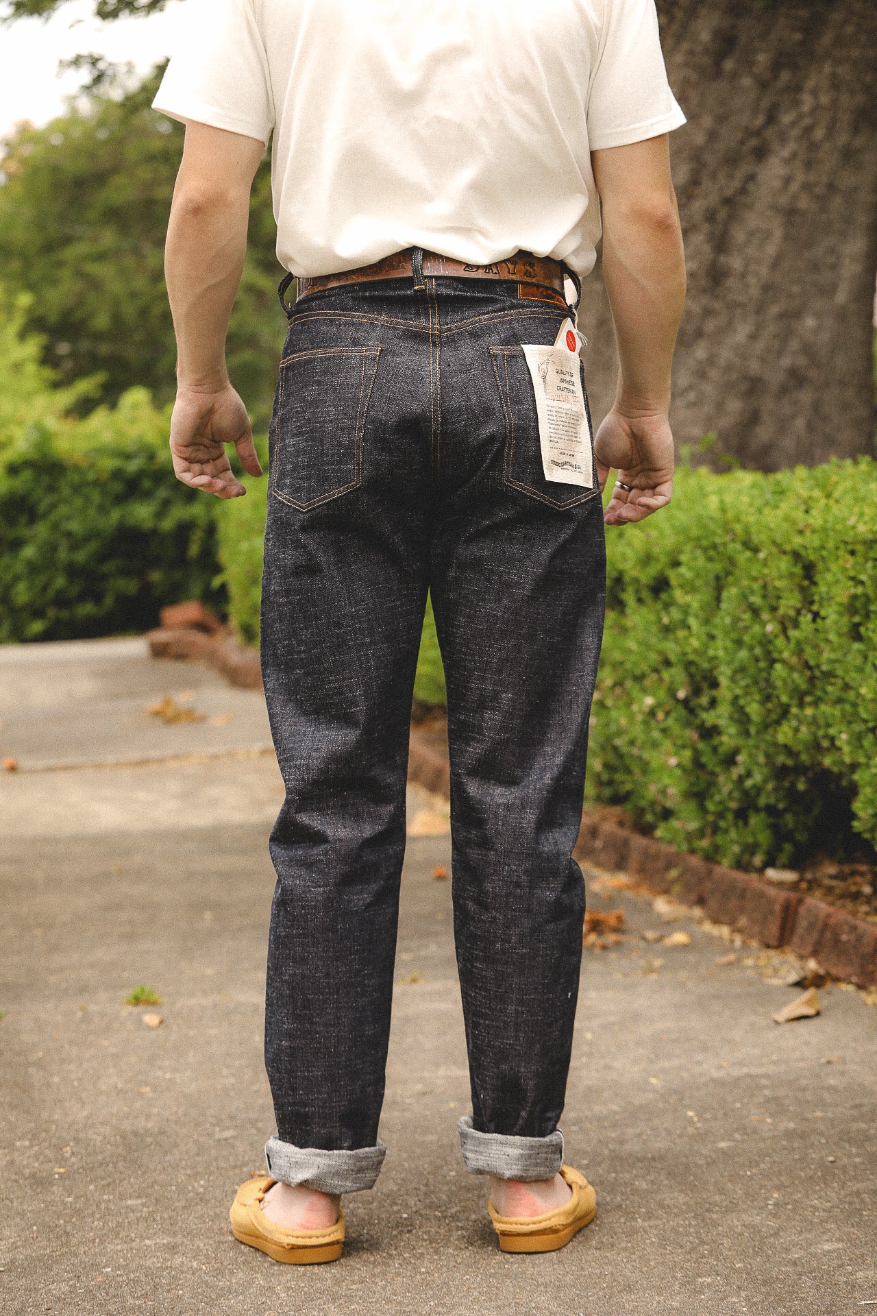 Person standing outdoors wearing dark denim jeans with rolled cuffs, a white t-shirt, and tan moccasin shoes near green bushes