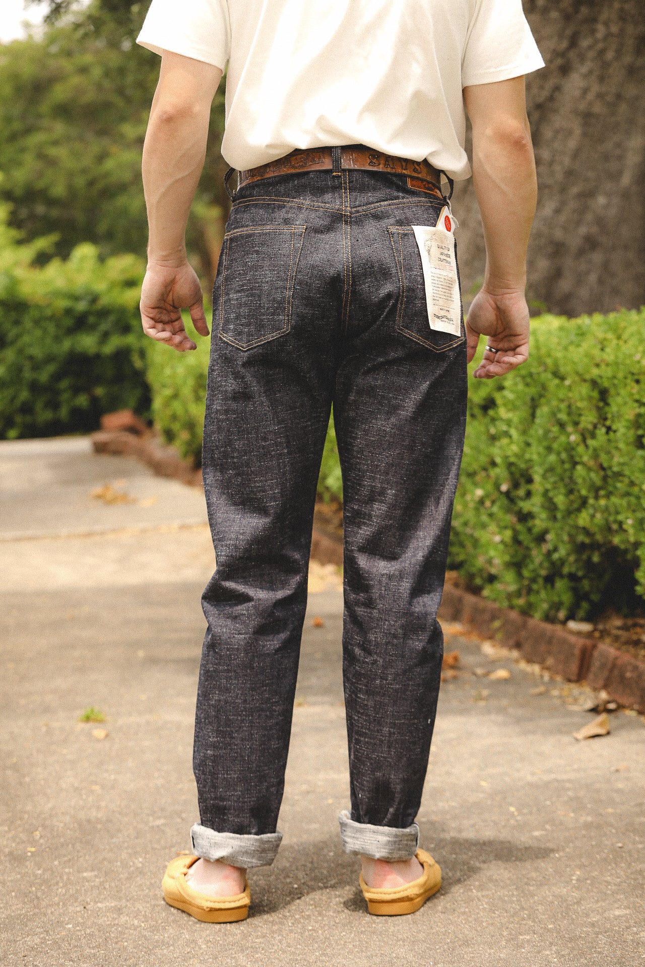 Rear view of person wearing dark raw denim jeans with rolled cuffs, white t-shirt, and brown belt outdoors