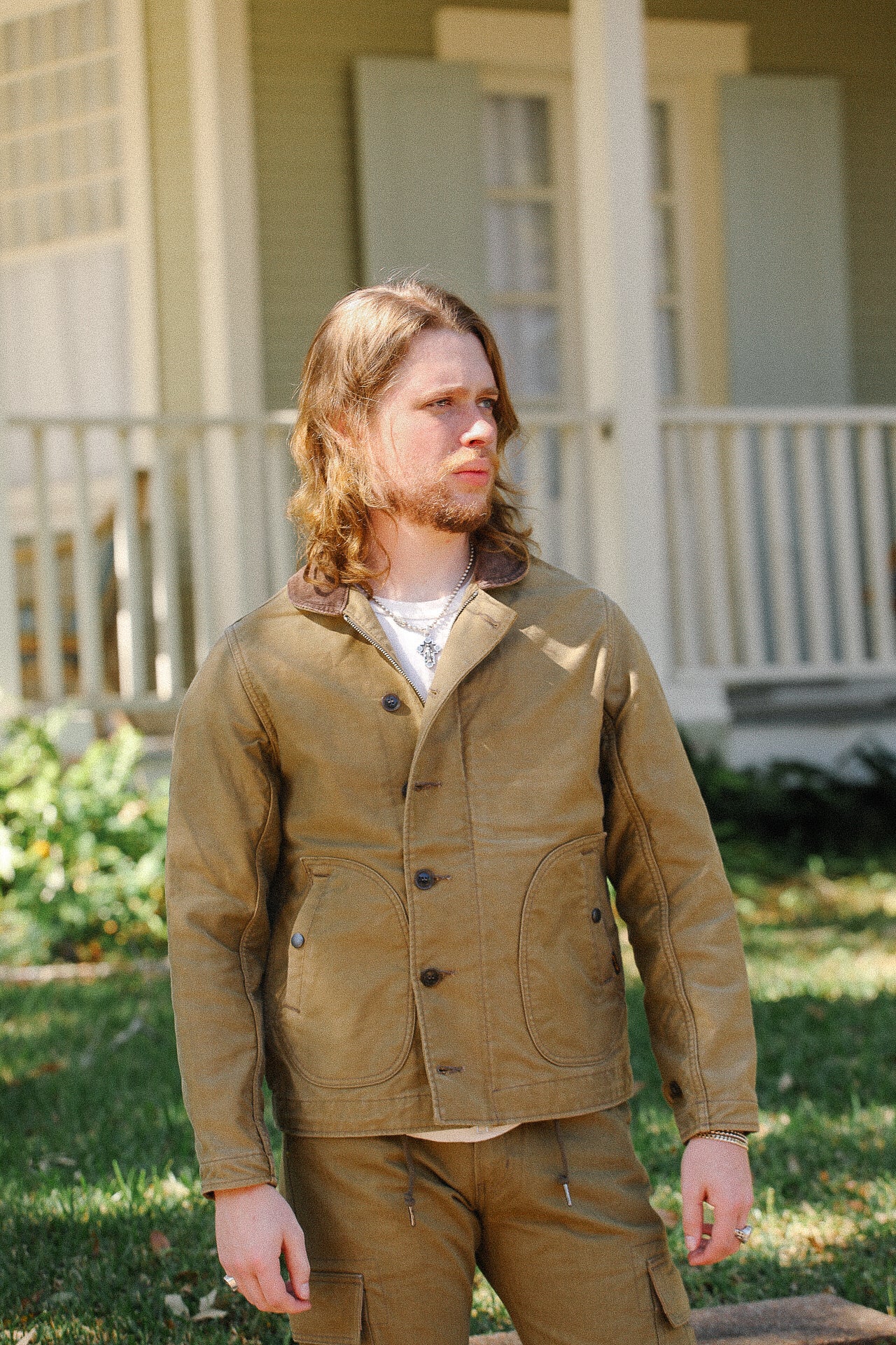 Man wearing brown rugged jacket and matching pants standing outdoors by a house porch
