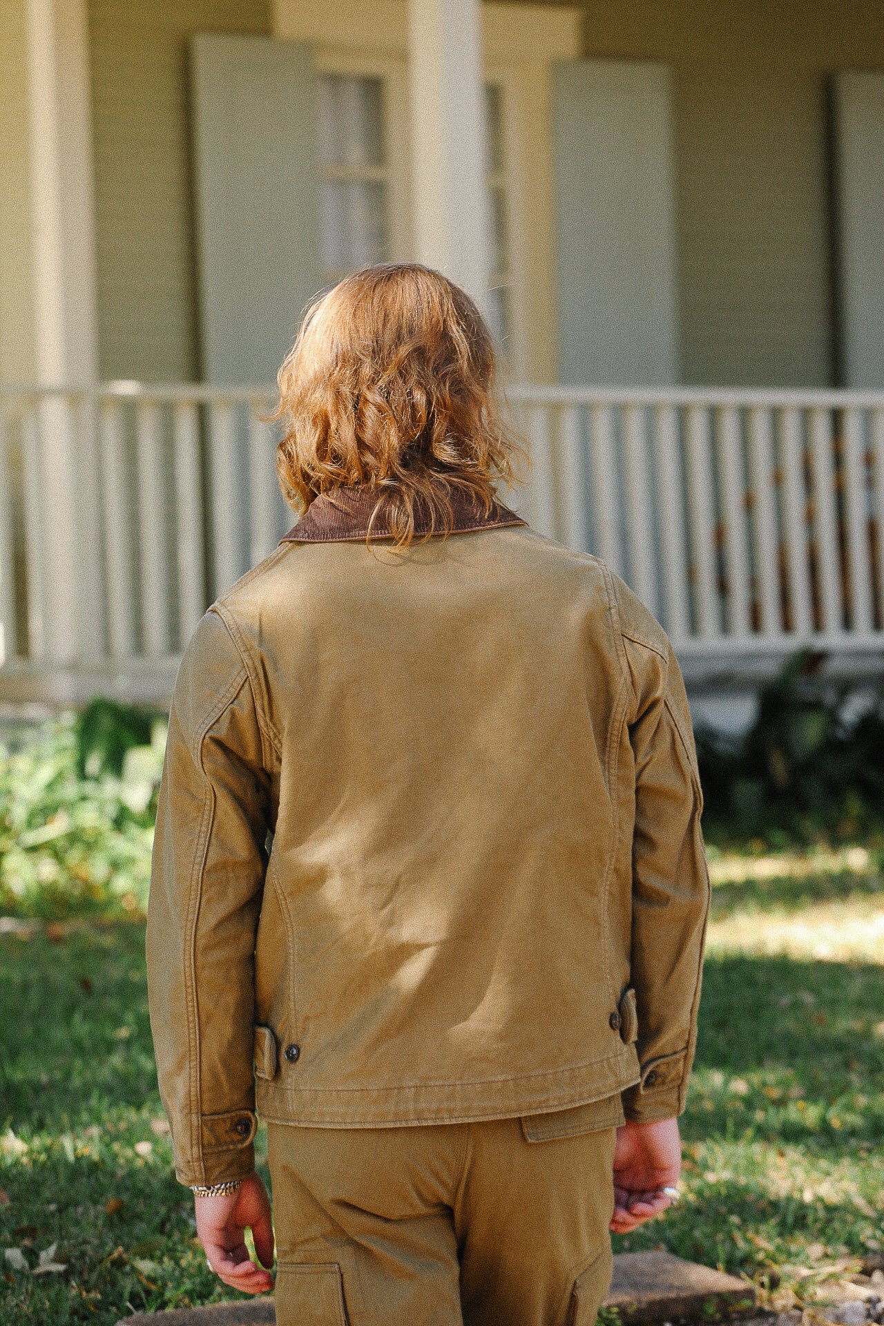 Person with shoulder-length hair wearing tan workwear jacket and pants standing outdoors in front of house porch