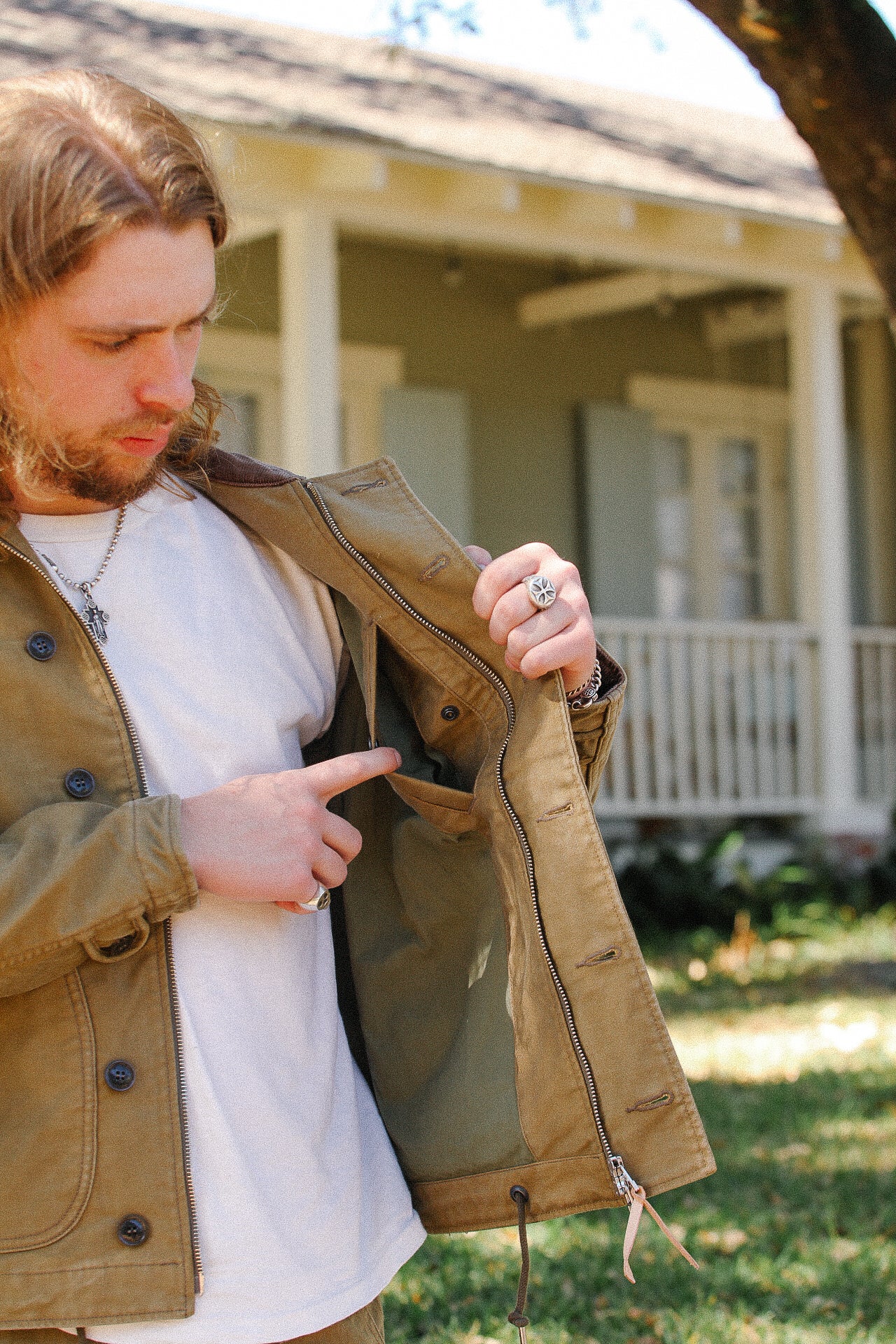 Man in tan jacket pointing to inside pocket outdoors by a porch with greenery