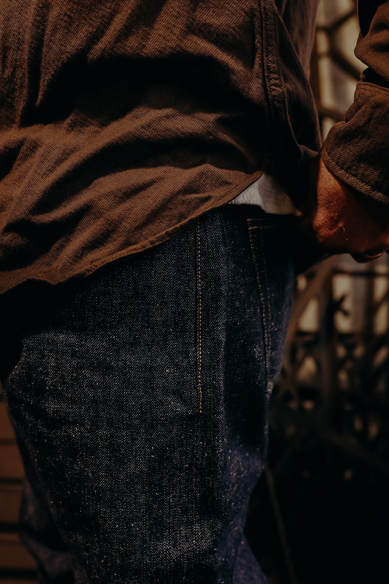 Close-up of a person wearing dark denim jeans and a brown textured shirt with a metal gate background