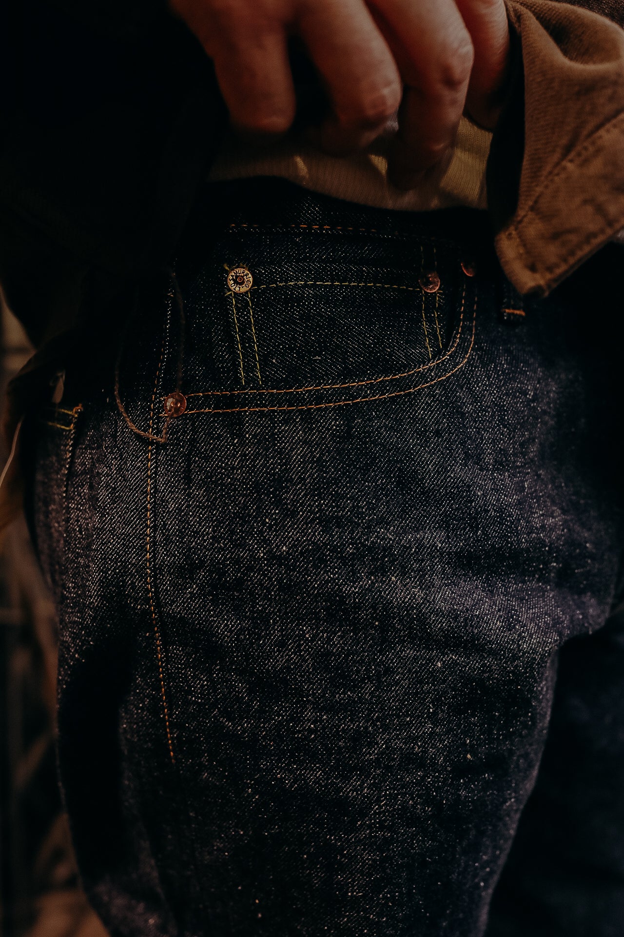 Close-up of dark raw denim jeans pocket with detailed stitching and copper rivets, hand adjusting waistband