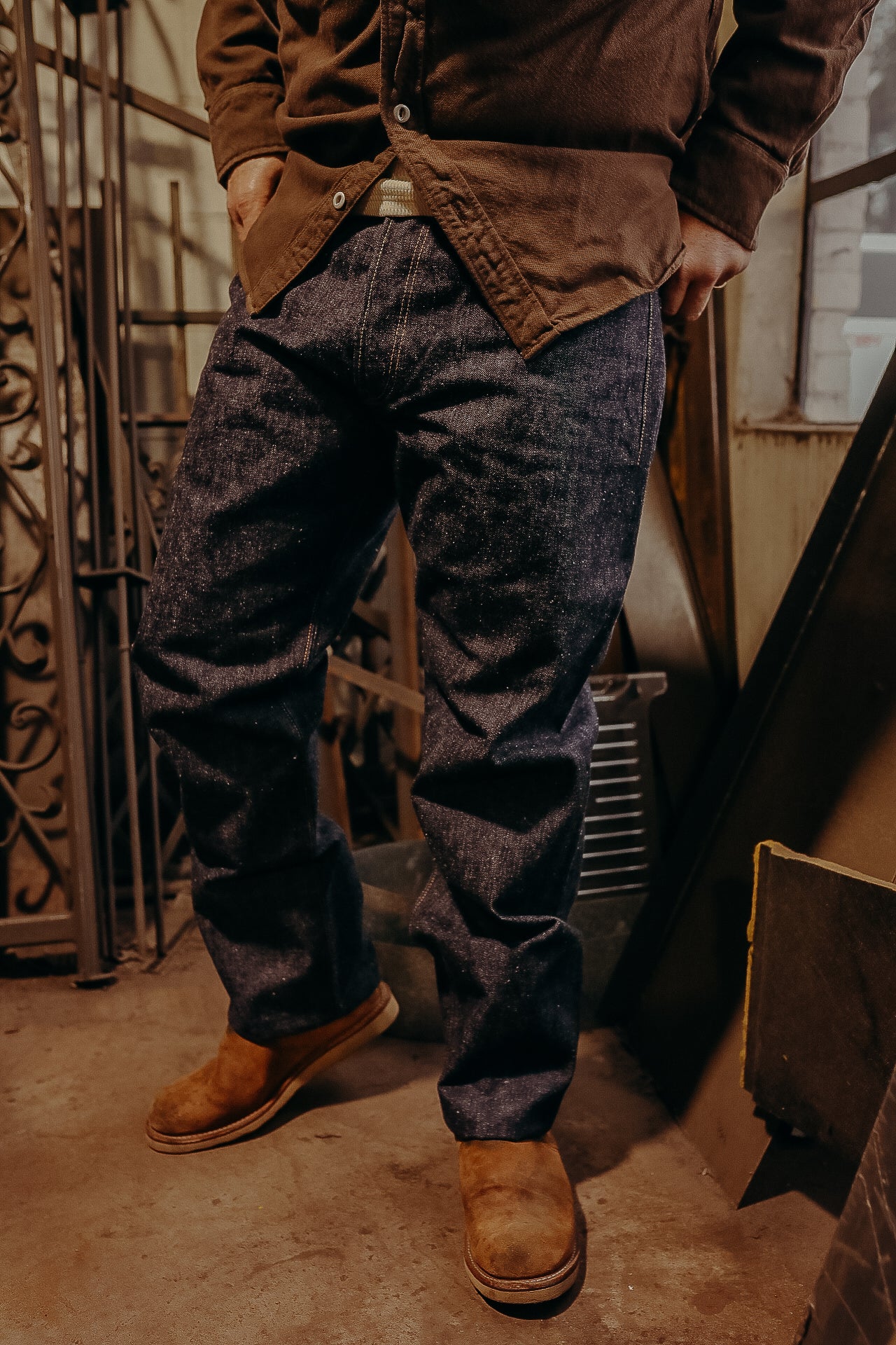 Close-up of person wearing dark denim jeans, brown jacket, and tan work boots in a workshop setting