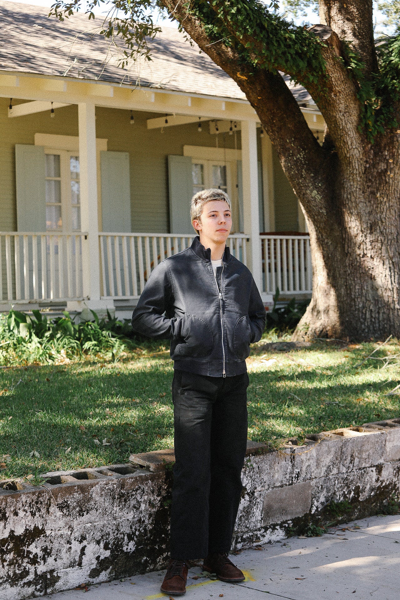 Person wearing black jacket and pants standing outside near a tree and porch of house
