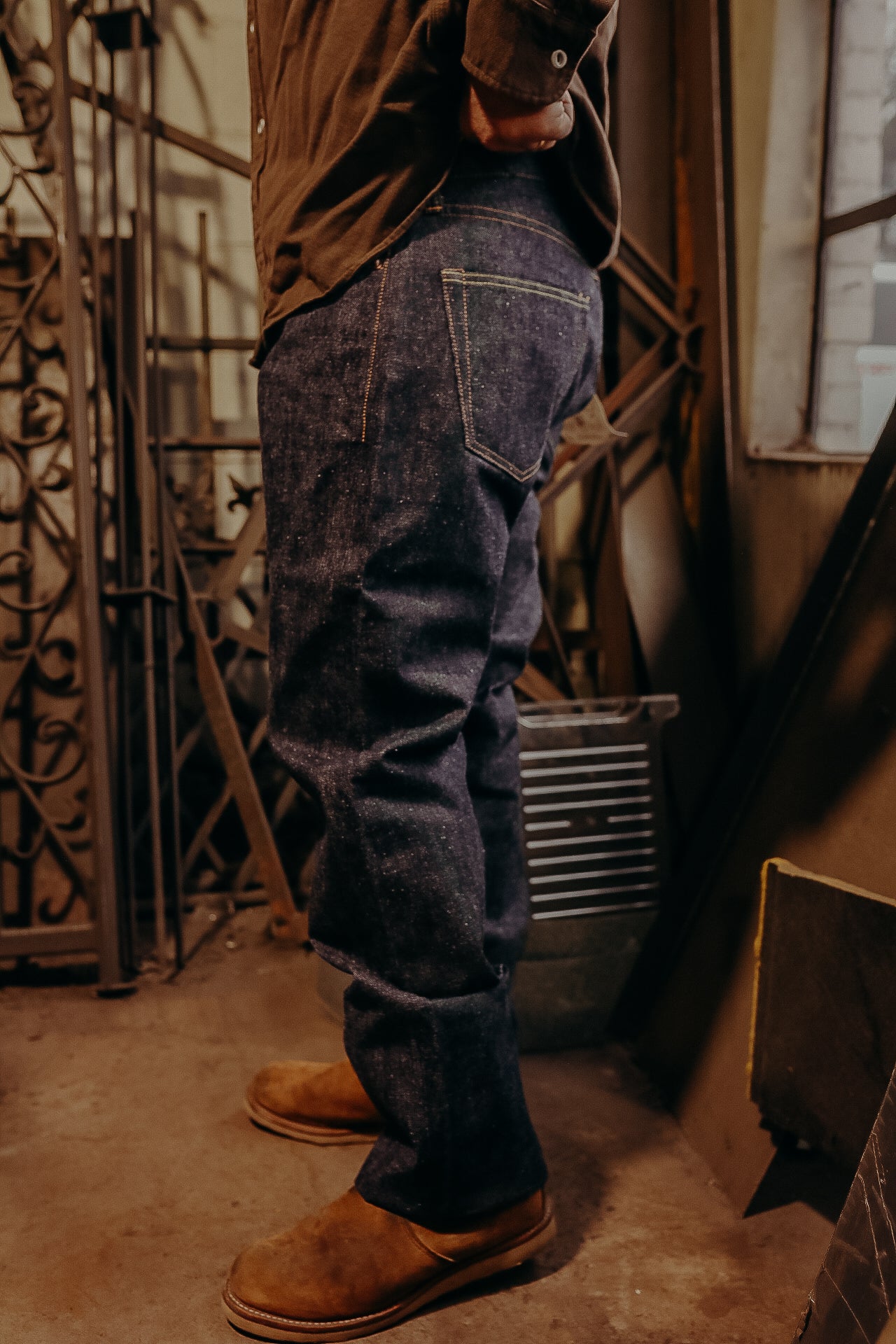 Person wearing dark blue raw denim jeans and brown leather boots in a rustic workshop with metalwork