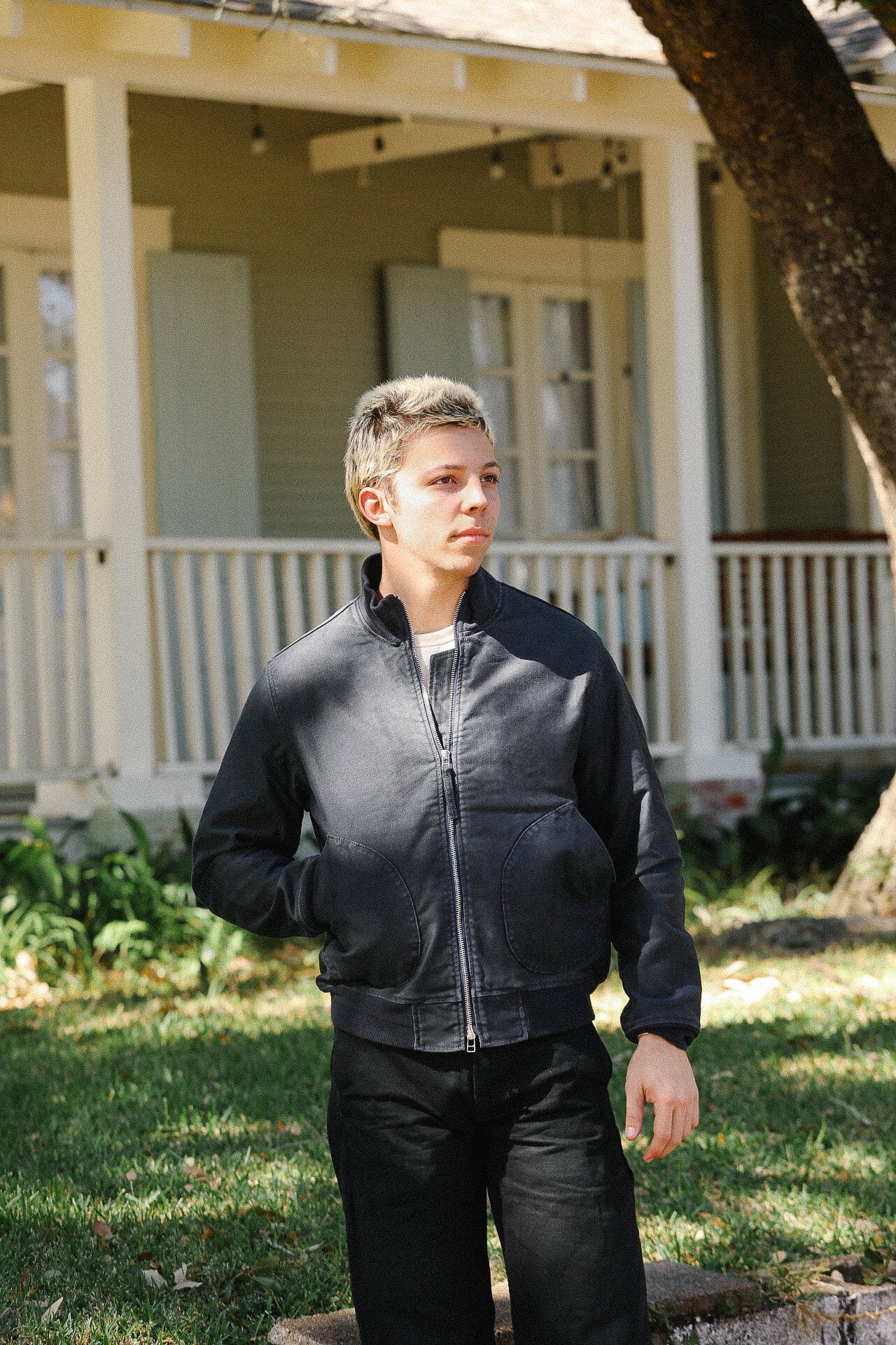 Young man wearing black leather jacket and pants standing outdoors in front of house porch in daylight