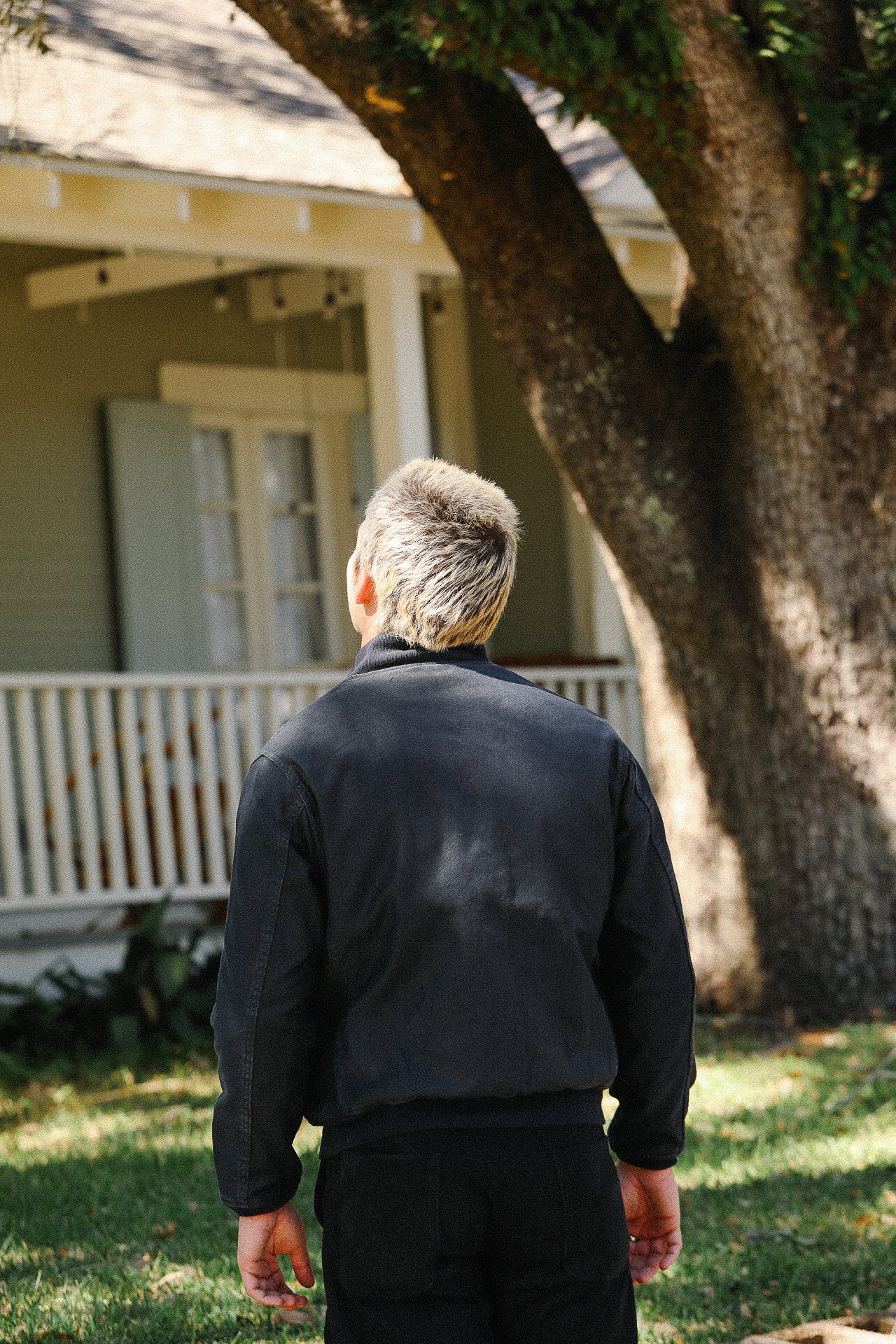 Person with short blonde hair wearing black jacket and pants standing outside near tree and porch