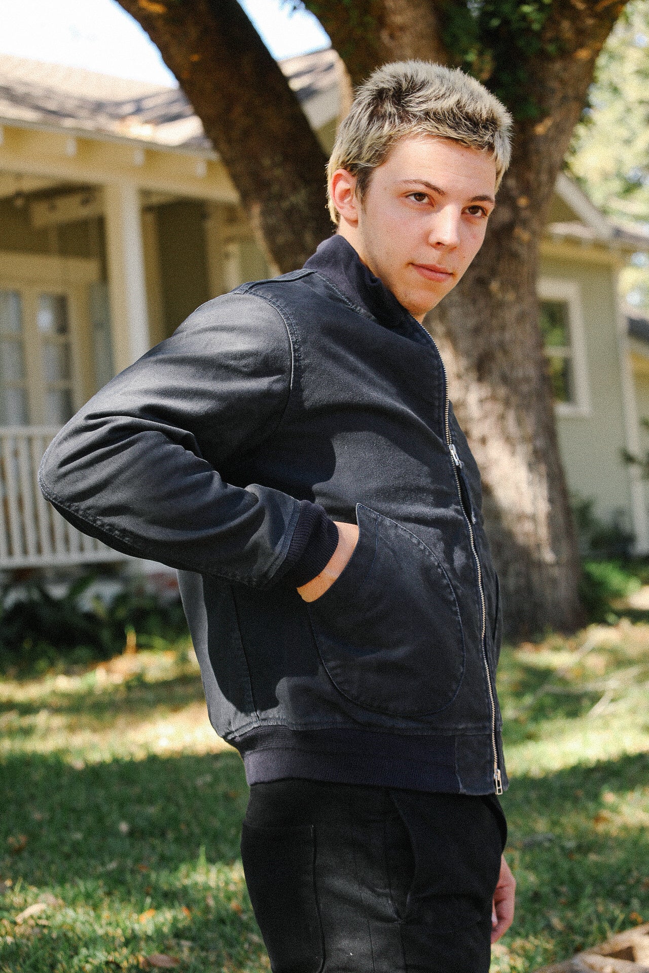Young man wearing a black zip-up jacket with hands in pockets standing outdoors near a tree and house