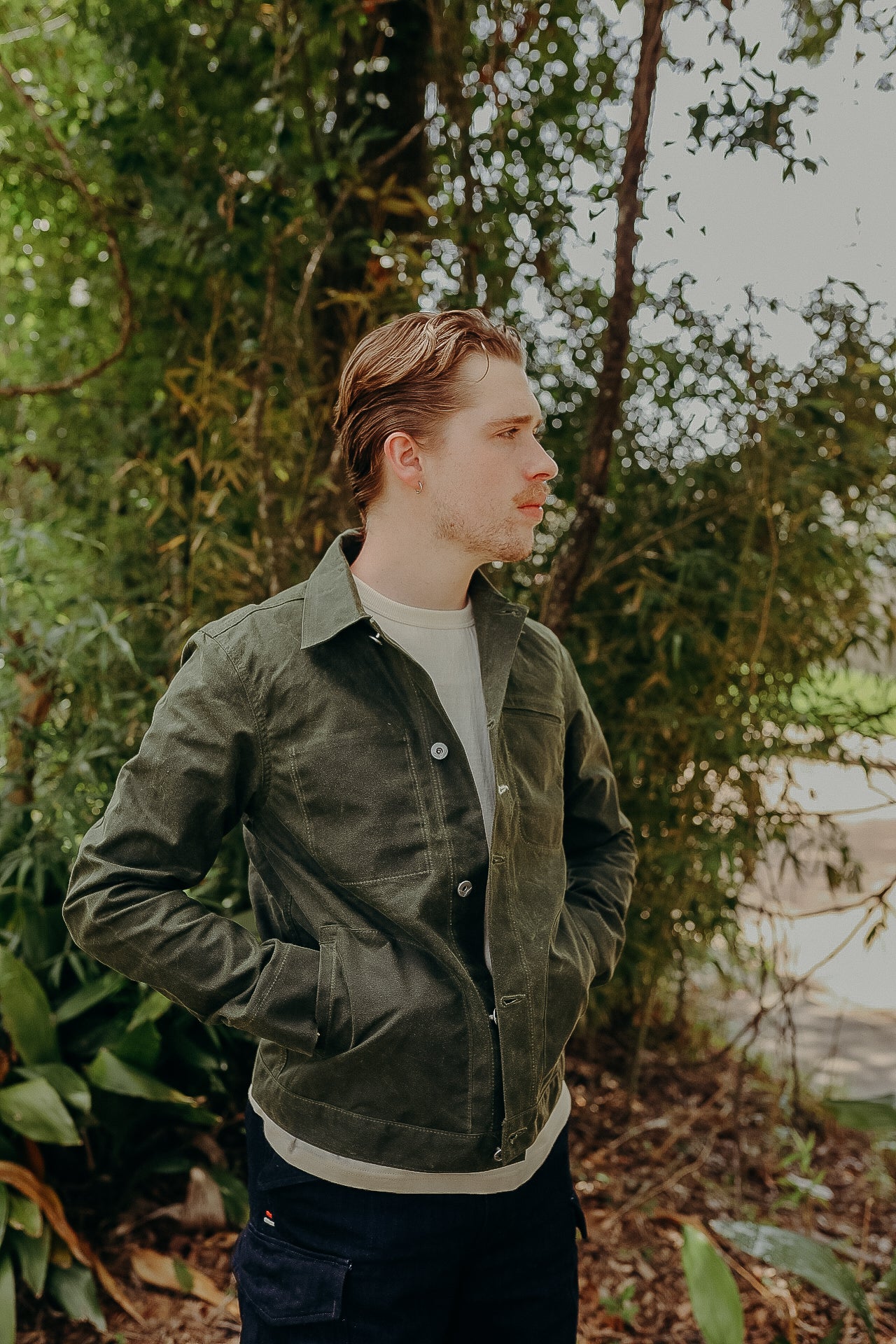 Man wearing olive green canvas chore jacket and white shirt outdoors with greenery background
