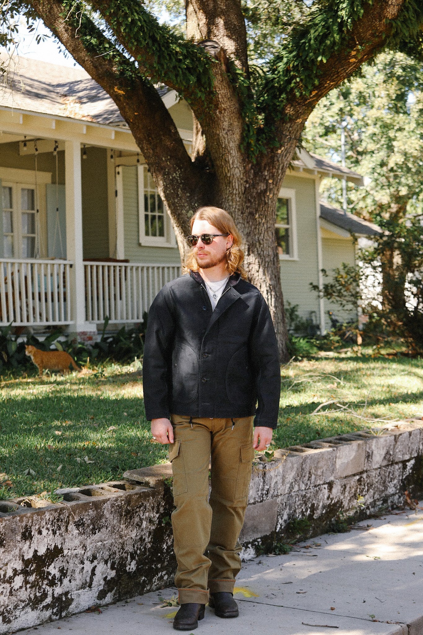 Man wearing black jacket, tan cargo pants, and boots standing by stone wall in front of a house with large tree