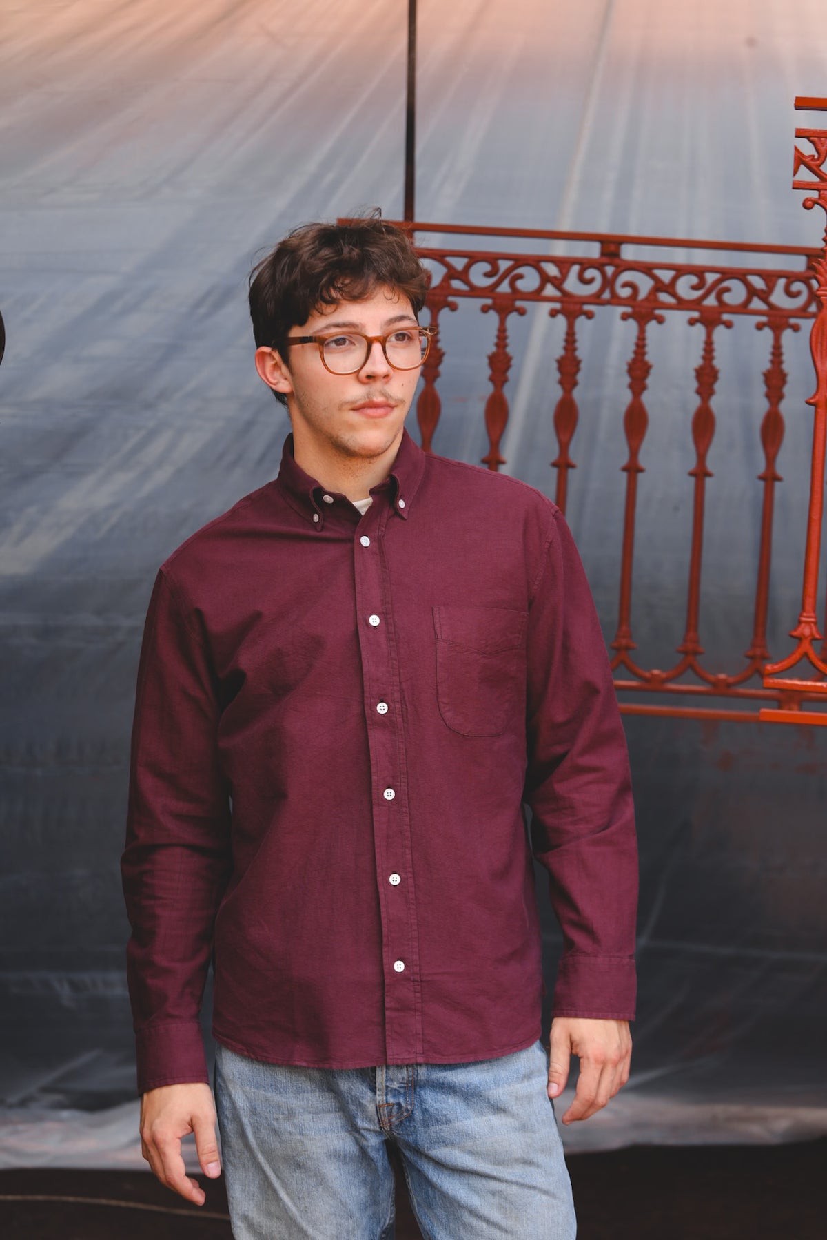 A young man wearing a burgundy long-sleeve button-down shirt and light blue jeans, standing in front of a gray draped backdrop with a red metal railing.
