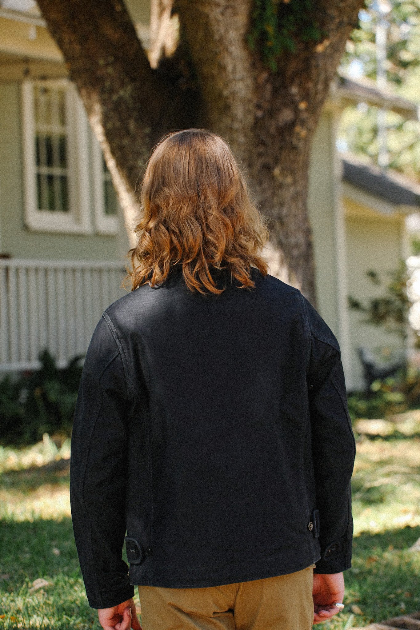 Person with shoulder-length hair wearing a black jacket and tan pants standing outdoors near a tree