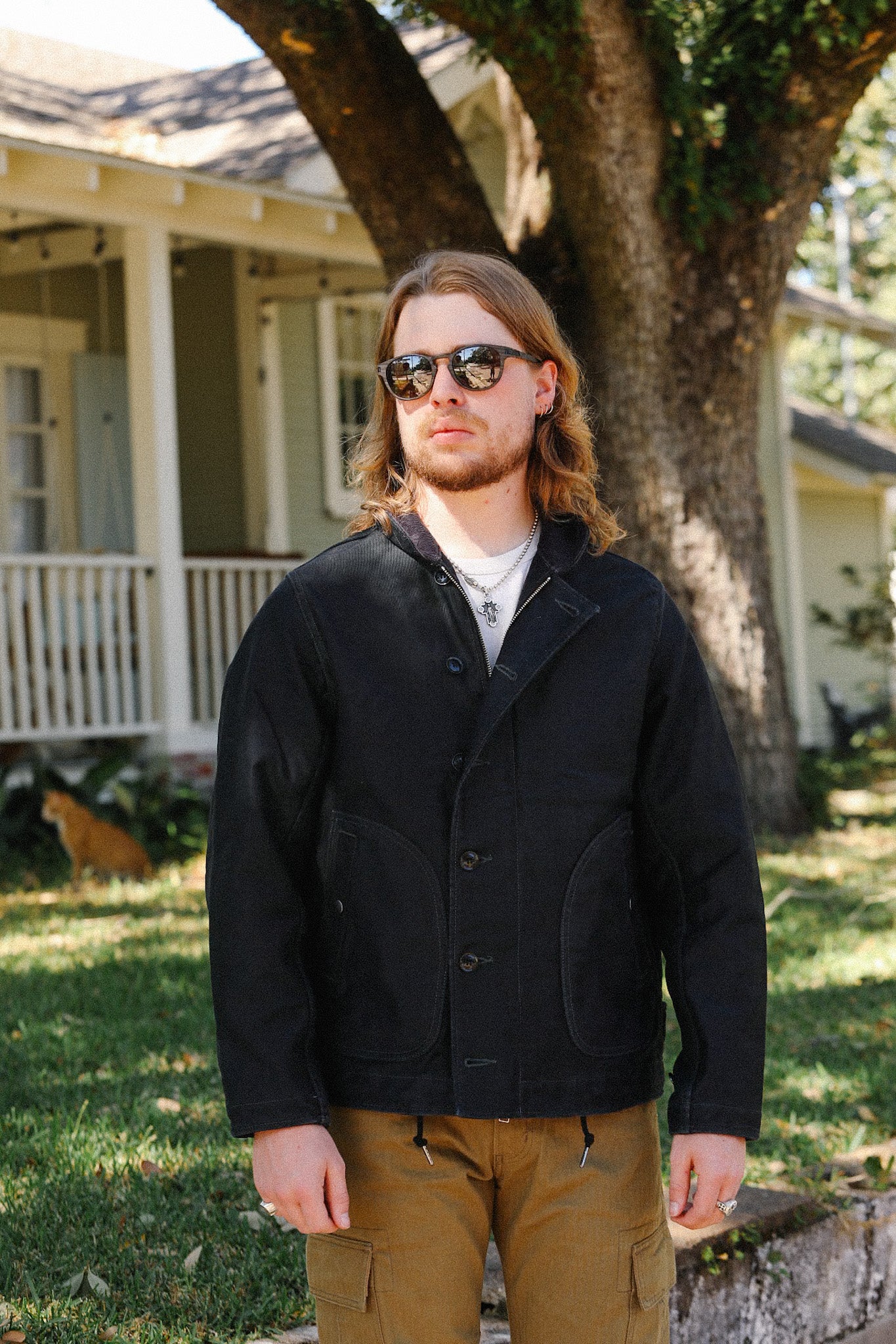 Man wearing black button-up jacket and sunglasses standing outdoors with house and tree in background